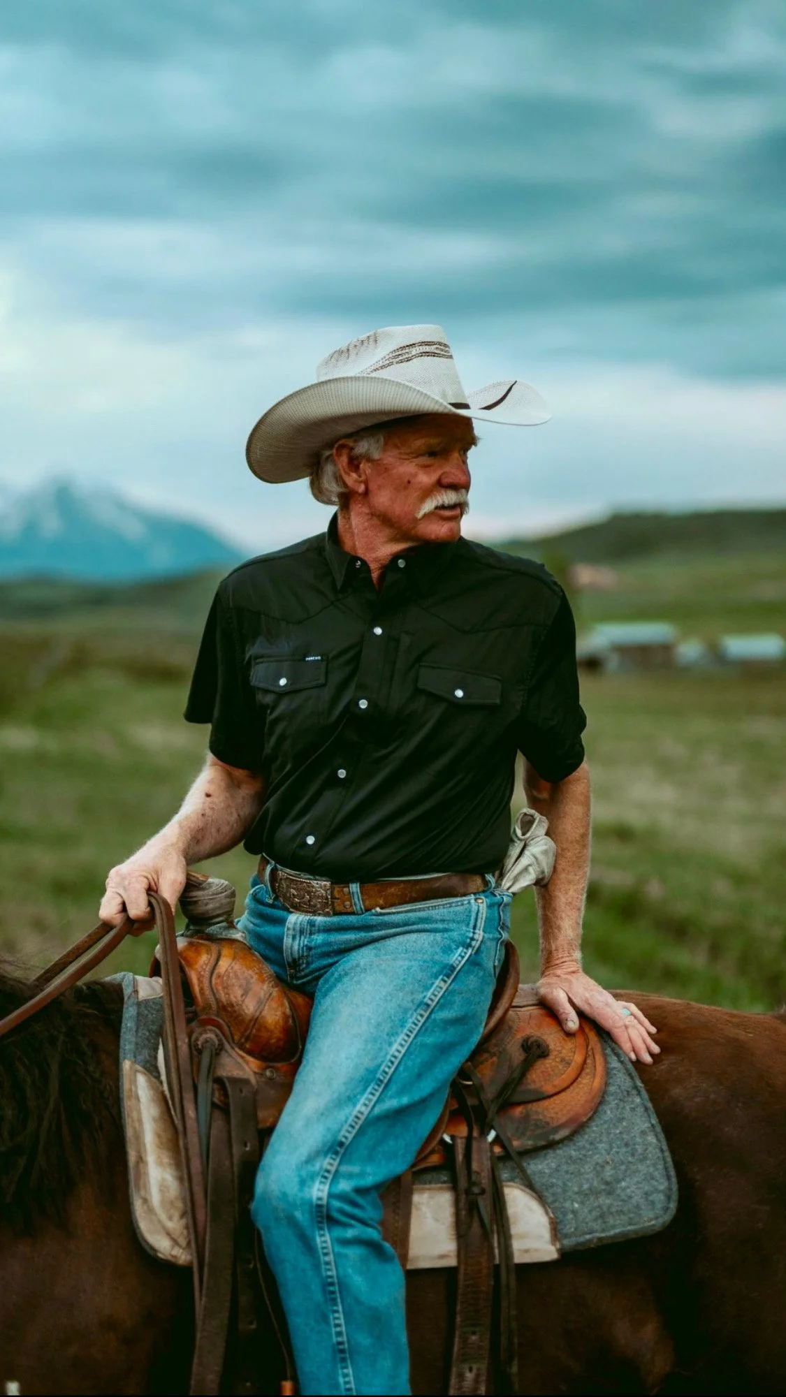 An older man with white hair and a mustache, wearing a cowboy hat, black shirt, blue jeans, and a belt, riding a horse outdoors with a scenic landscape of green fields, mountains, and a cloudy sky in the background.