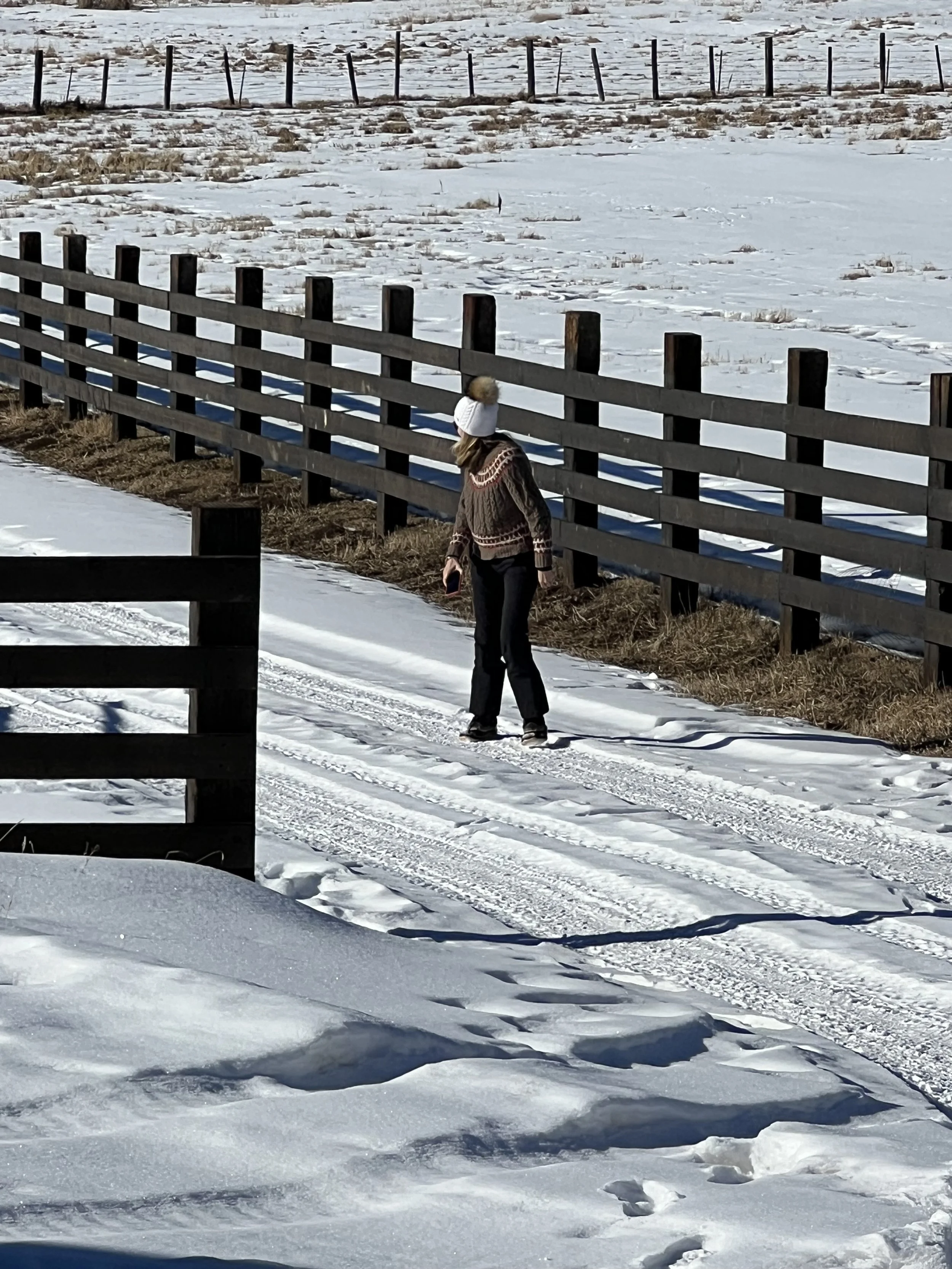 A person walking on a snow-covered path next to a wooden fence in a snowy landscape, wearing a white winter hat with a pom-pom and a patterned sweater.