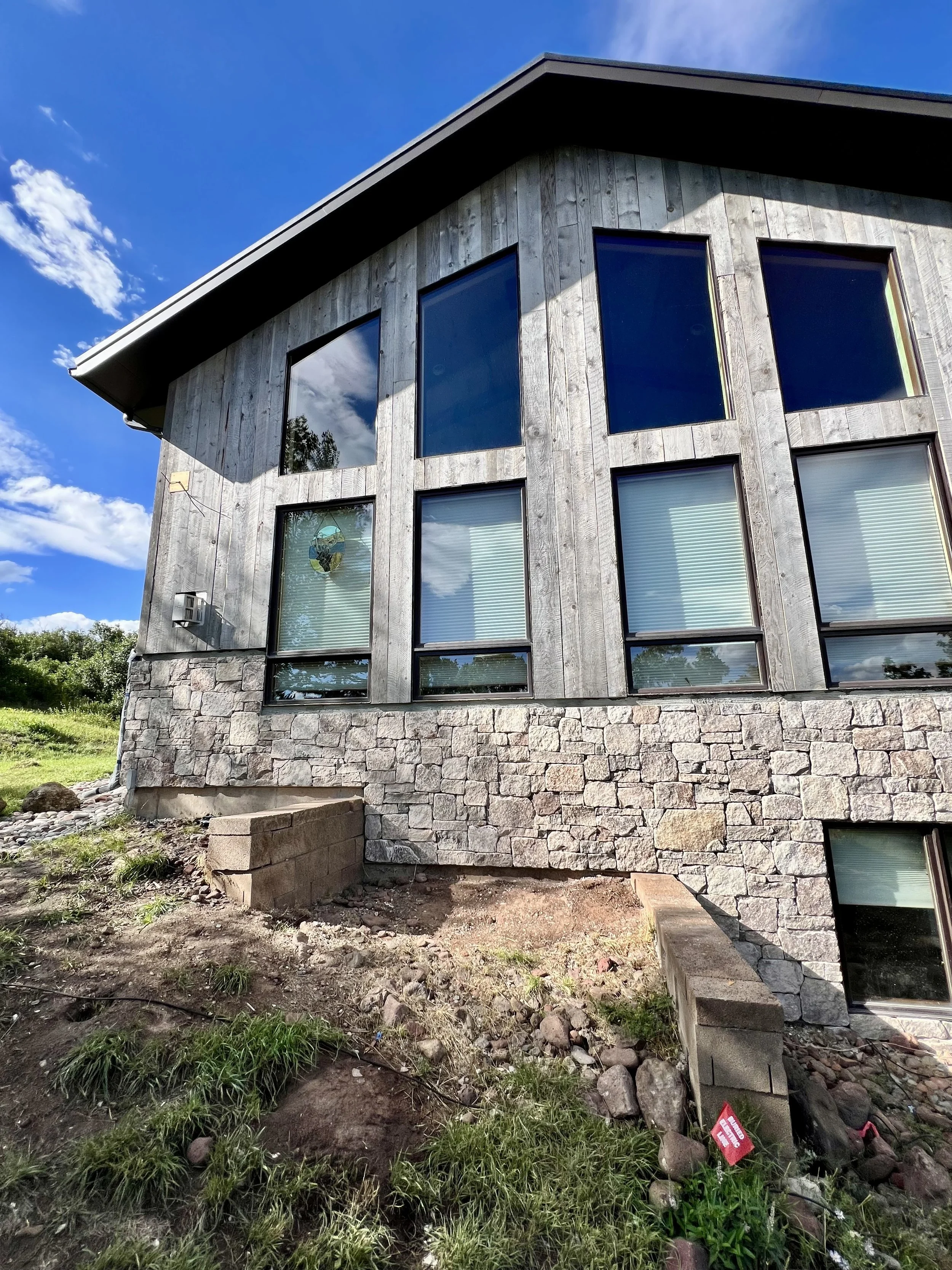 Exterior of a modern house with large windows, wooden siding, stone foundation, and a blue sky with clouds.