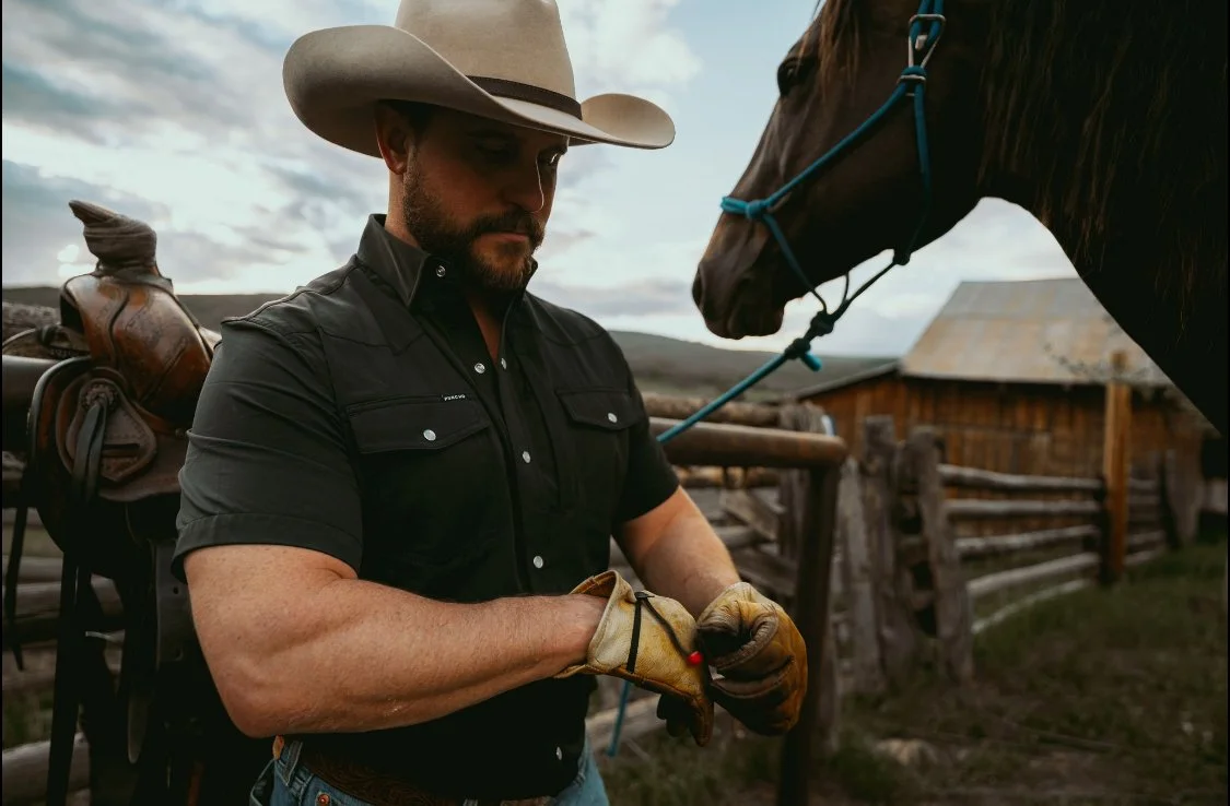 A man wearing a cowboy hat and black shirt prepares to ride a horse on a farm with a wooden barn in the background.