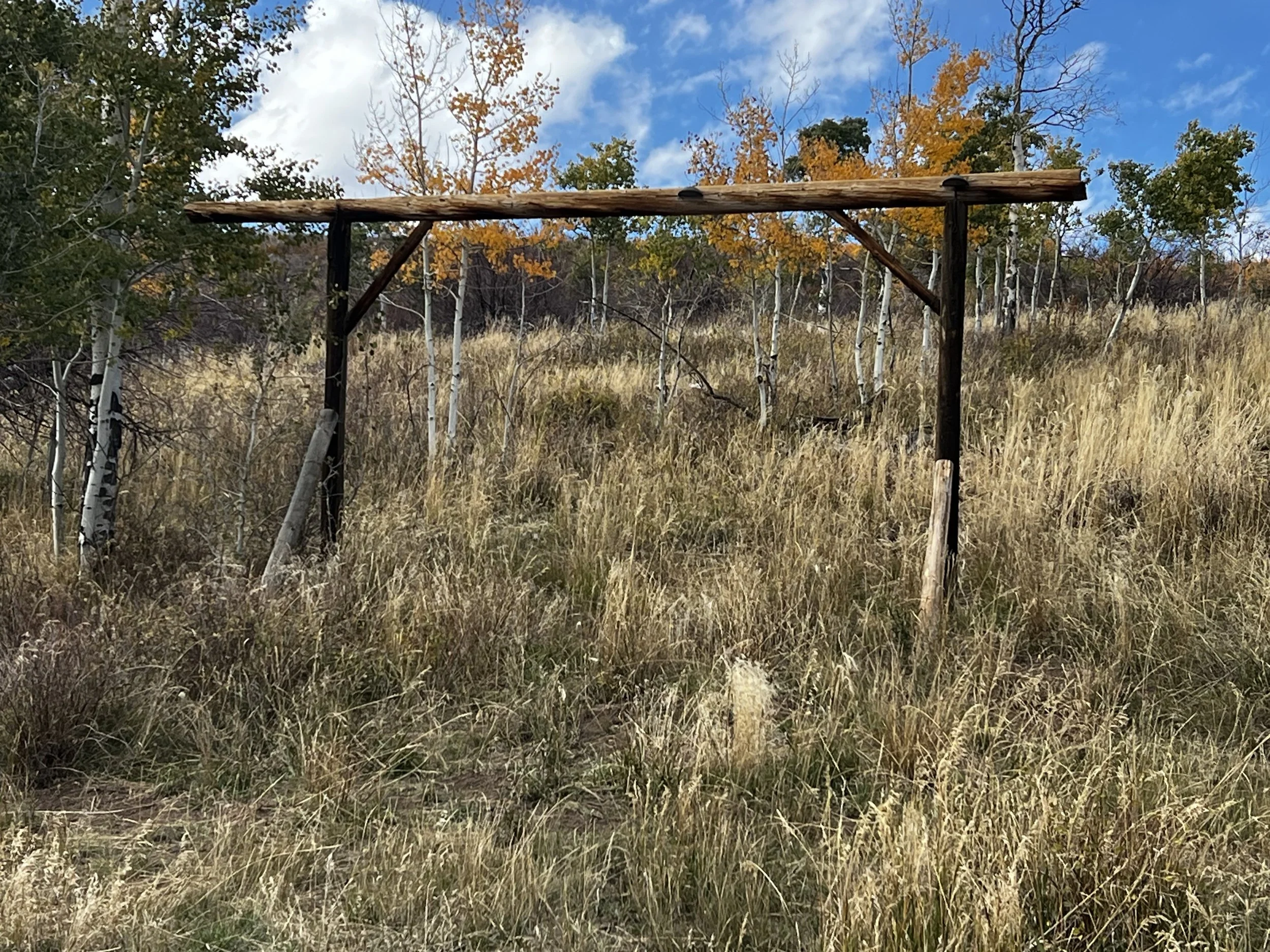 A rustic wooden archway on a grassy hillside with trees showing fall foliage and a partly cloudy blue sky.