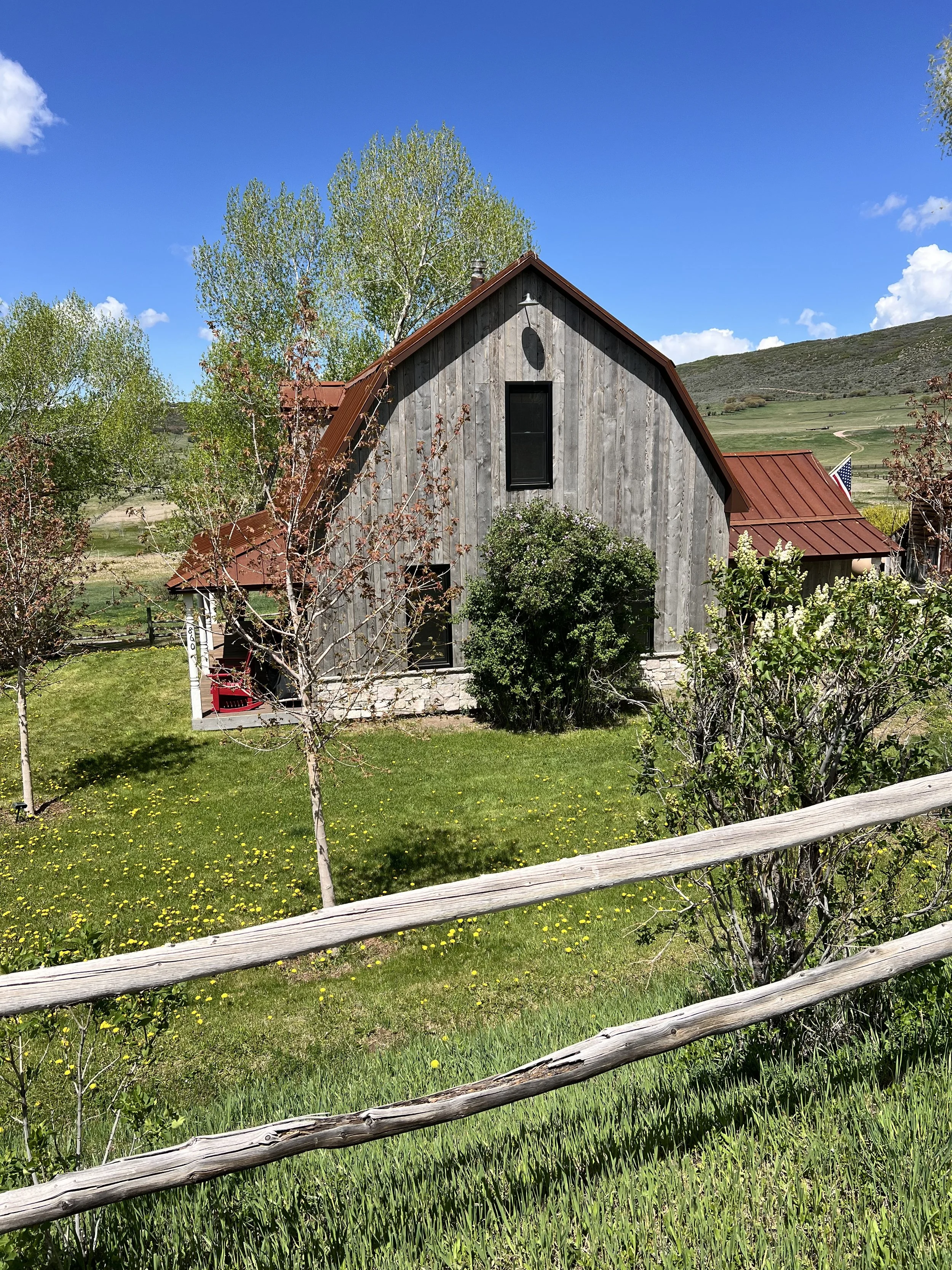 A rustic barn with weathered wood siding and a red metal roof surrounded by trees and a green lawn, with a clear blue sky overhead.