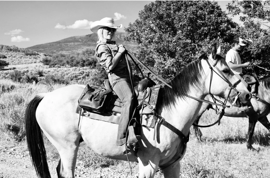 A woman riding a white horse outdoors, wearing a cowboy hat and sunglasses, with a scenic landscape and rolling hills in the background.