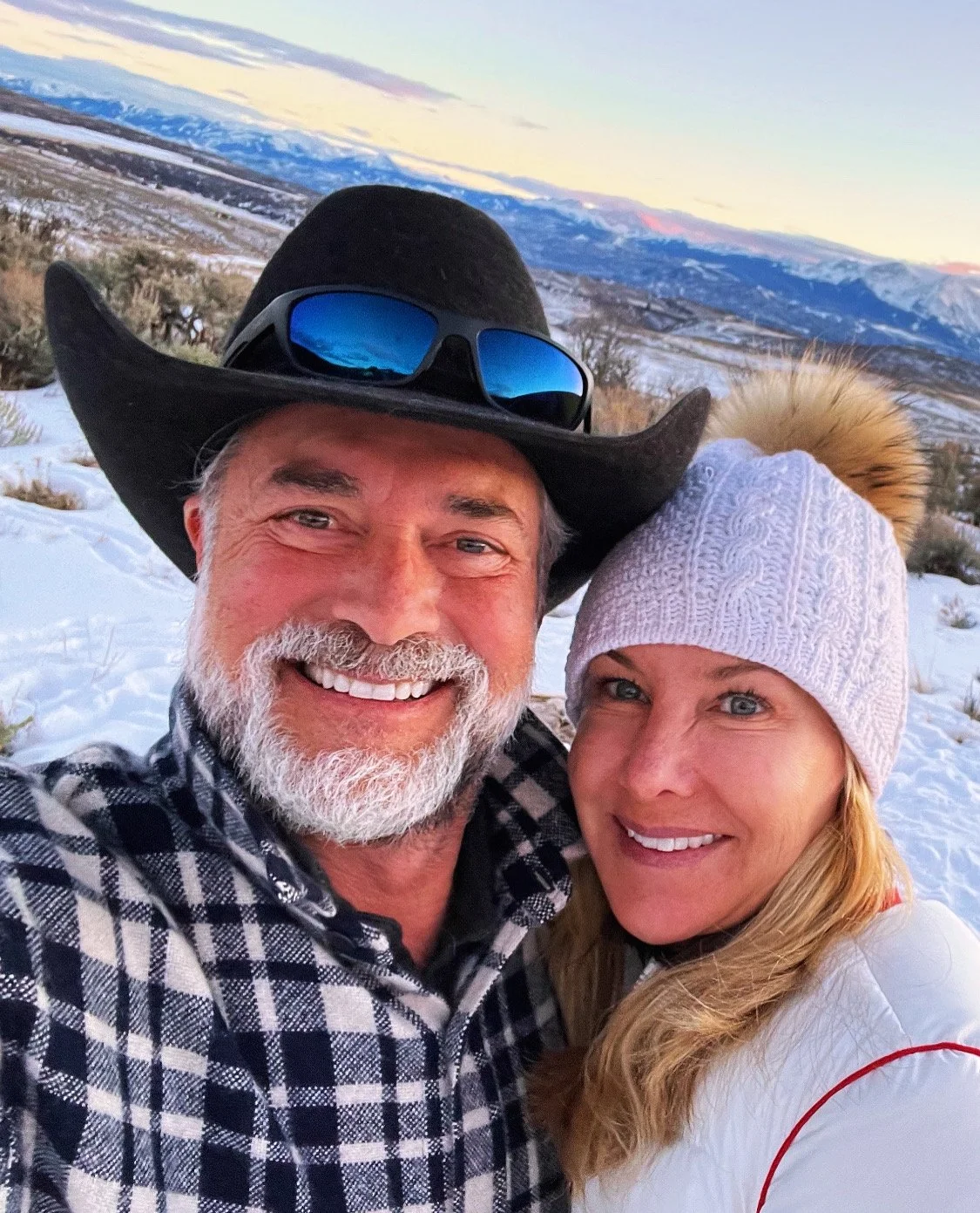 A smiling couple taking a selfie outdoors in a snowy mountainous landscape during sunset. The man is wearing a black cowboy hat, sunglasses, and a black-and-white checkered shirt. The woman is wearing a white knit hat with a fur pompom and a white ja
