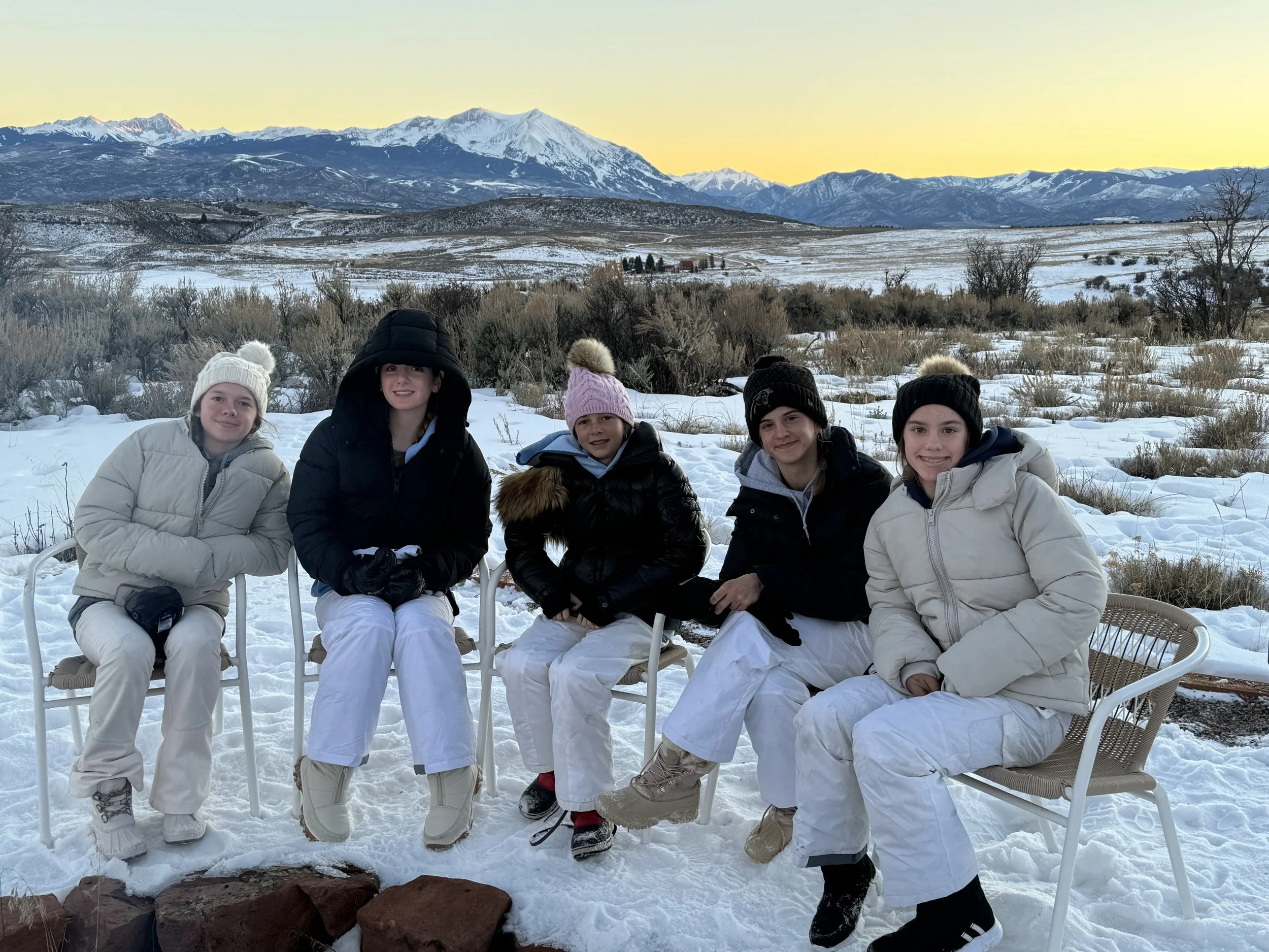 Five children dressed in winter clothing sitting on chairs outdoors in a snow-covered landscape with mountains in the background during sunset.