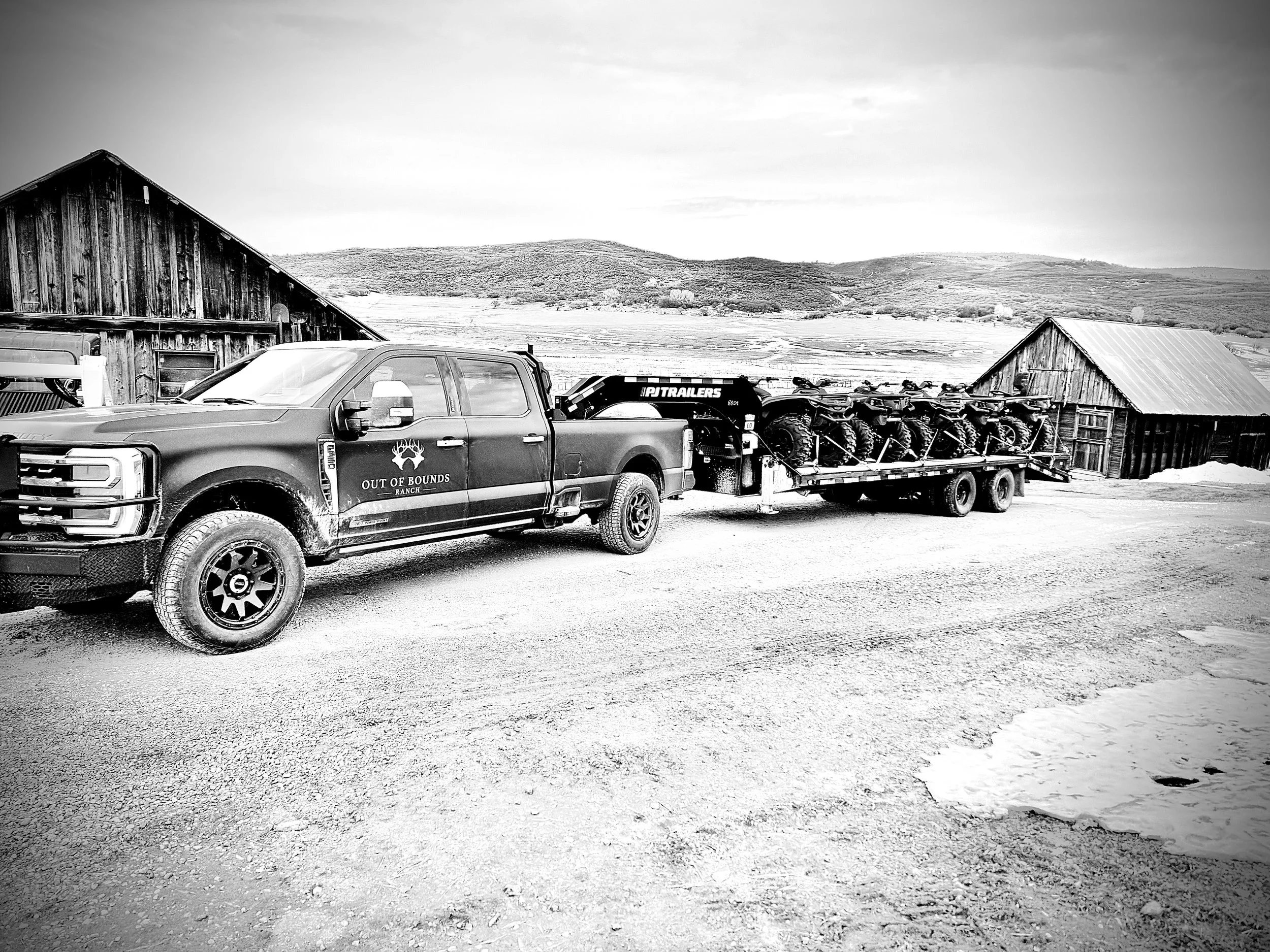 A pickup truck with a trailer carrying four all-terrain vehicles parked on a gravel road next to two rustic barns and hilly landscape in the background, captured in black and white.