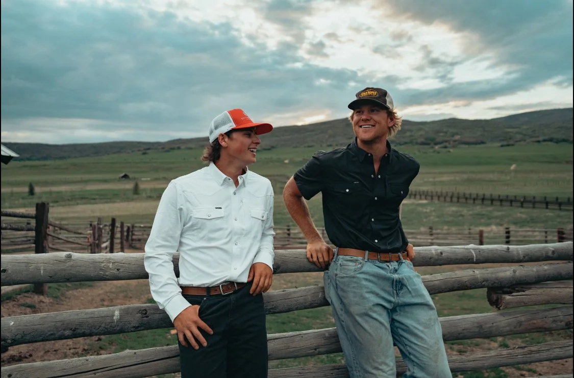 Two young men are standing outdoors by a wooden fence, smiling and talking to each other. One is wearing a white shirt and a red cap, while the other is wearing a black shirt and a black cap. The background features open fields, a cloudy sky, and rol