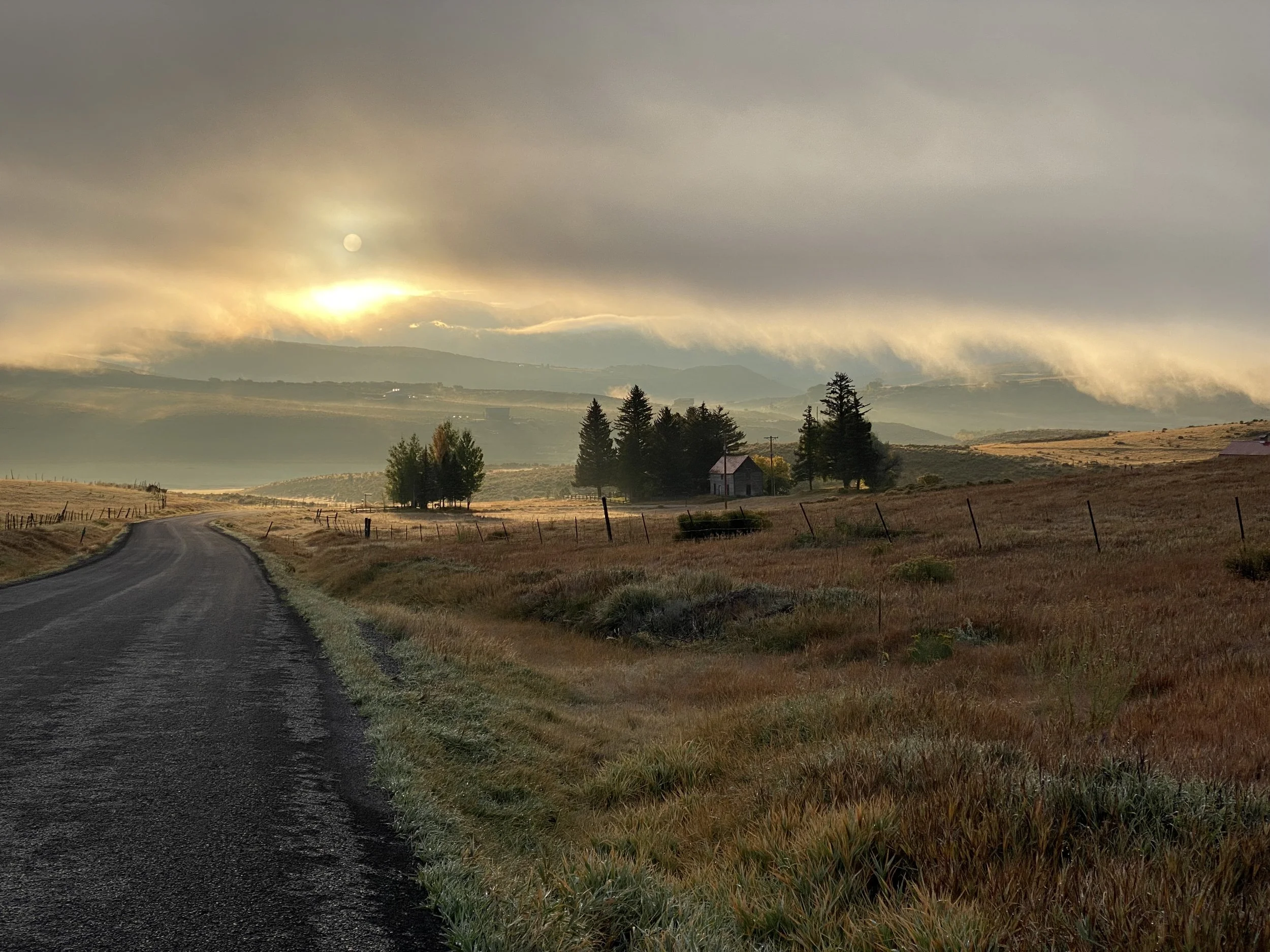 A rural landscape with a winding road, a small house, trees, and rolling hills under a cloudy sky with the sun partially obscured.