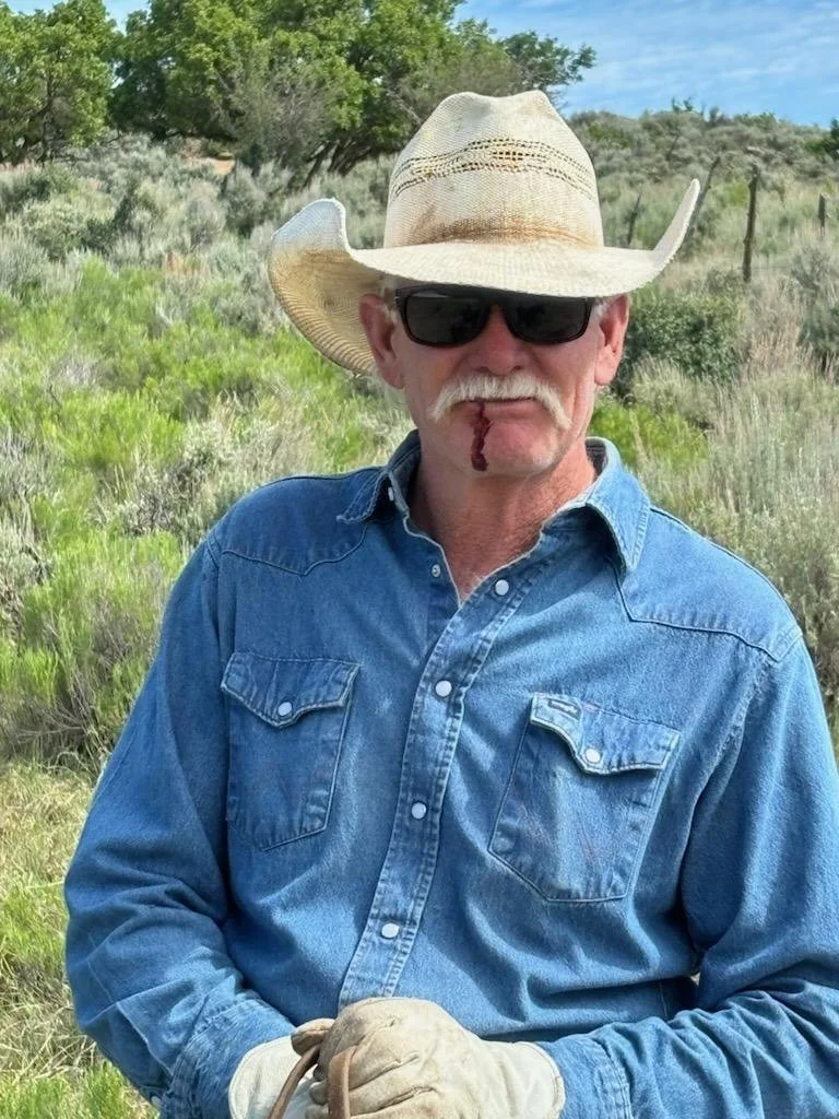 An older man wearing a cowboy hat, sunglasses, and a denim shirt standing outdoors in a grassy, bushy area with trees in the background.
