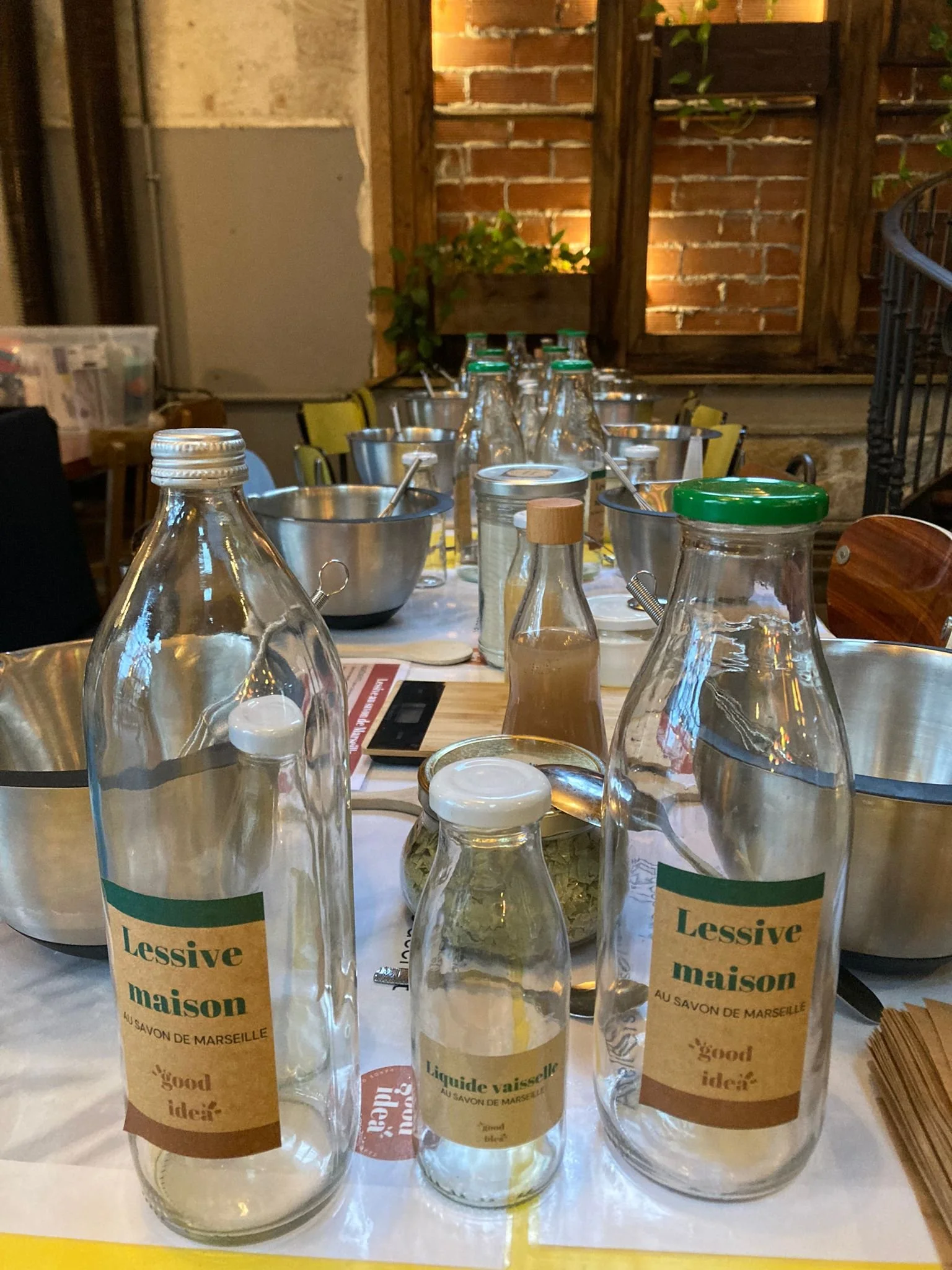 Bouteilles en verre étiquetées "Lessive maison" et "Liquide vaisselle", disposées sur une table avec des bols en métal et des ustensiles sous un éclairage chaleureux.