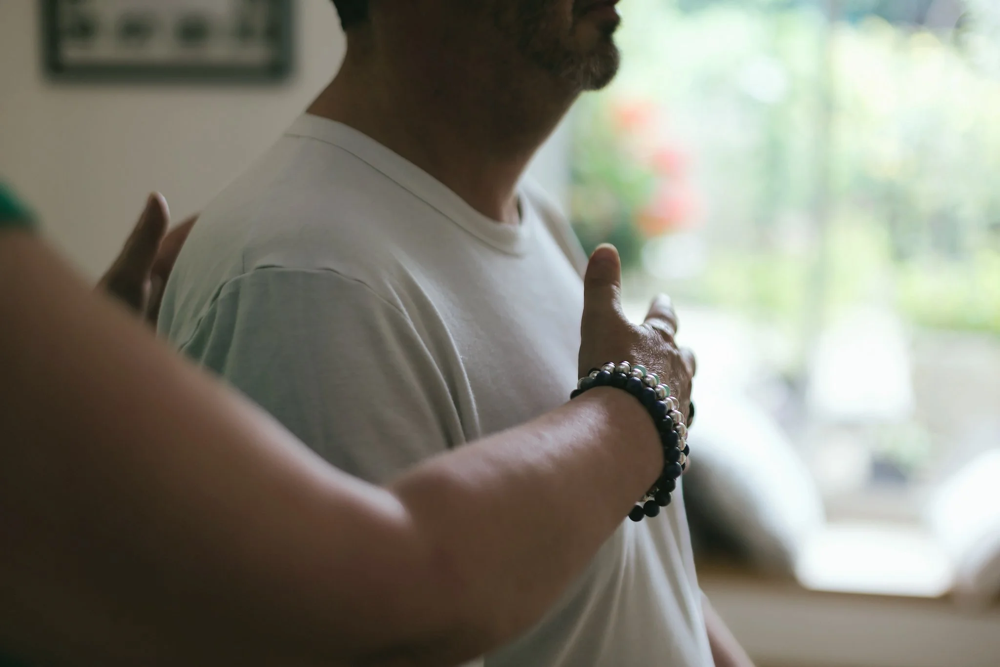 Un homme pratique la méditation ou la prière, touchant sa poitrine avec une main, portant un bracelet de perles, à côté d'une fenêtre avec un fond flou de nature.