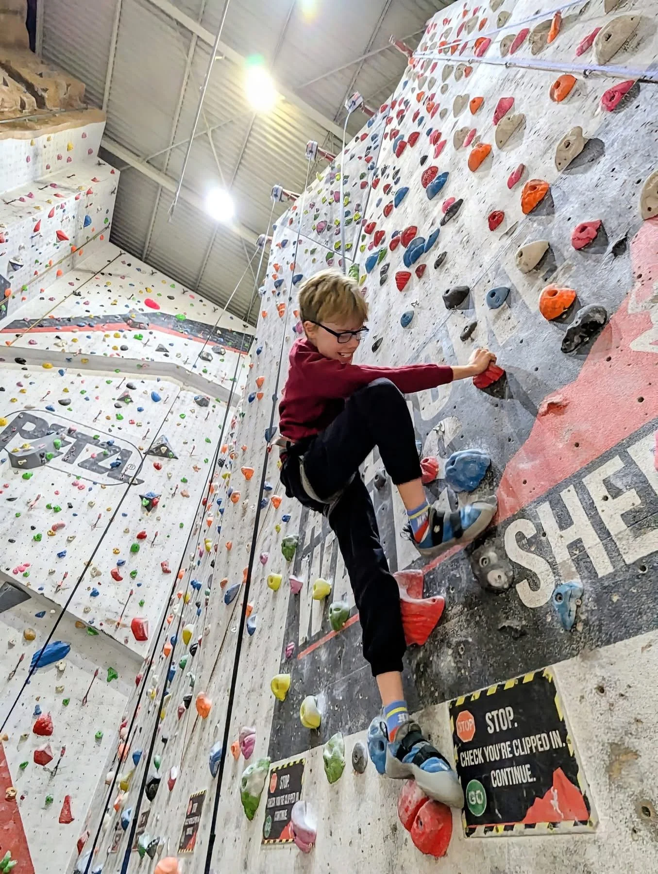 A person climbing an indoor rock wall with colorful holds, wearing glasses, a maroon hoodie, and black pants.