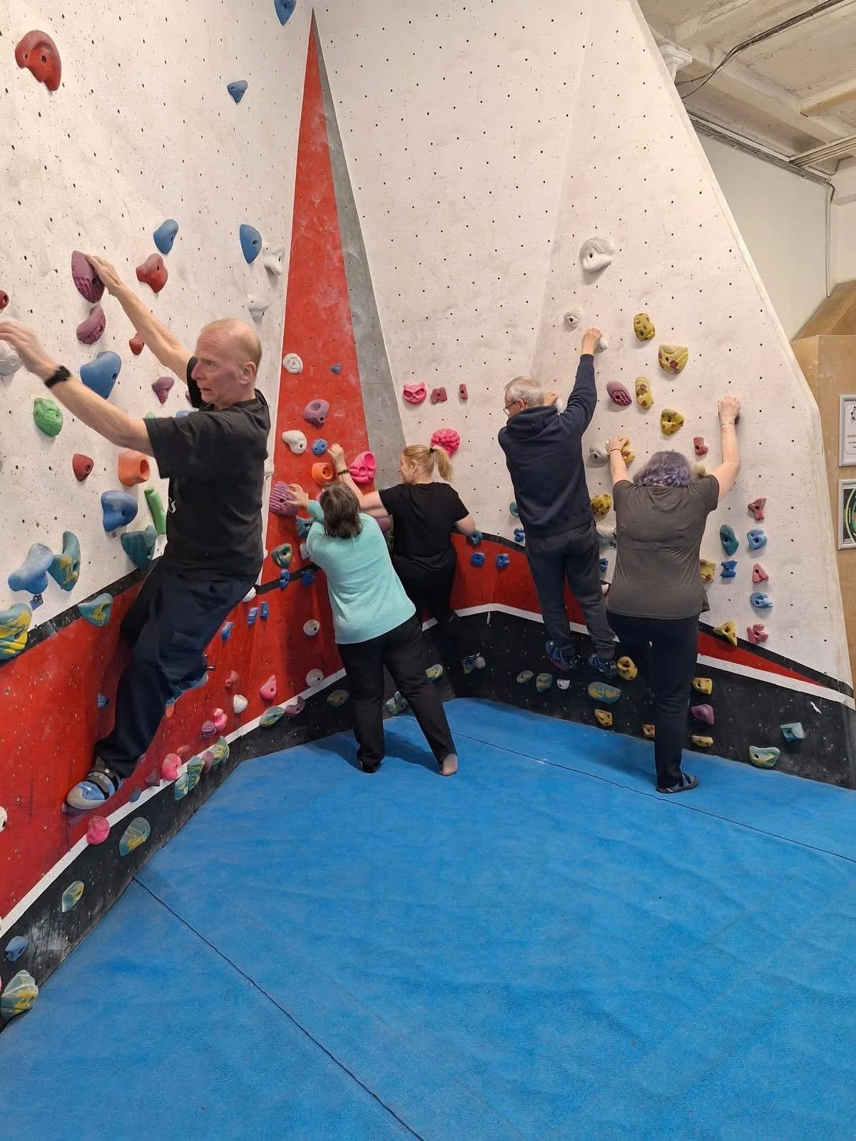 Five people climbing on an indoor bouldering wall with colorful holds, with a padded blue floor area.