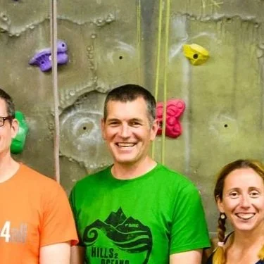 Smiling man in a green T-shirt standing in front of an indoor rock climbing wall with colorful holds.