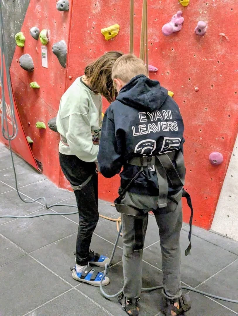 Two kids with climbing harnesses preparing to climb an indoor rock climbing wall with red and gray sections, colorful holds, and climbing equipment on the floor.
