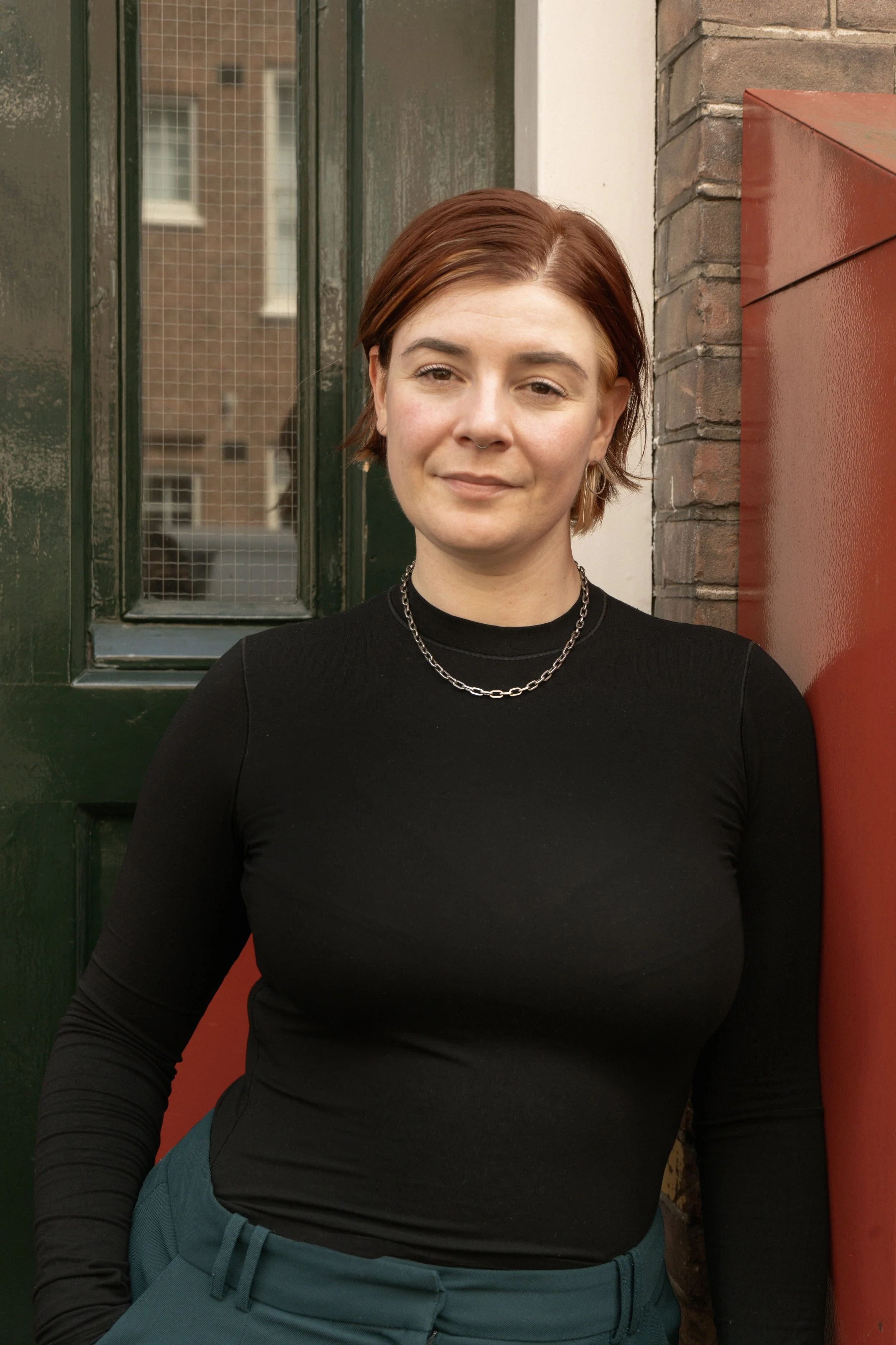 A woman with short red hair wearing a black long-sleeve top and teal pants standing outside against a brick wall and green door.
