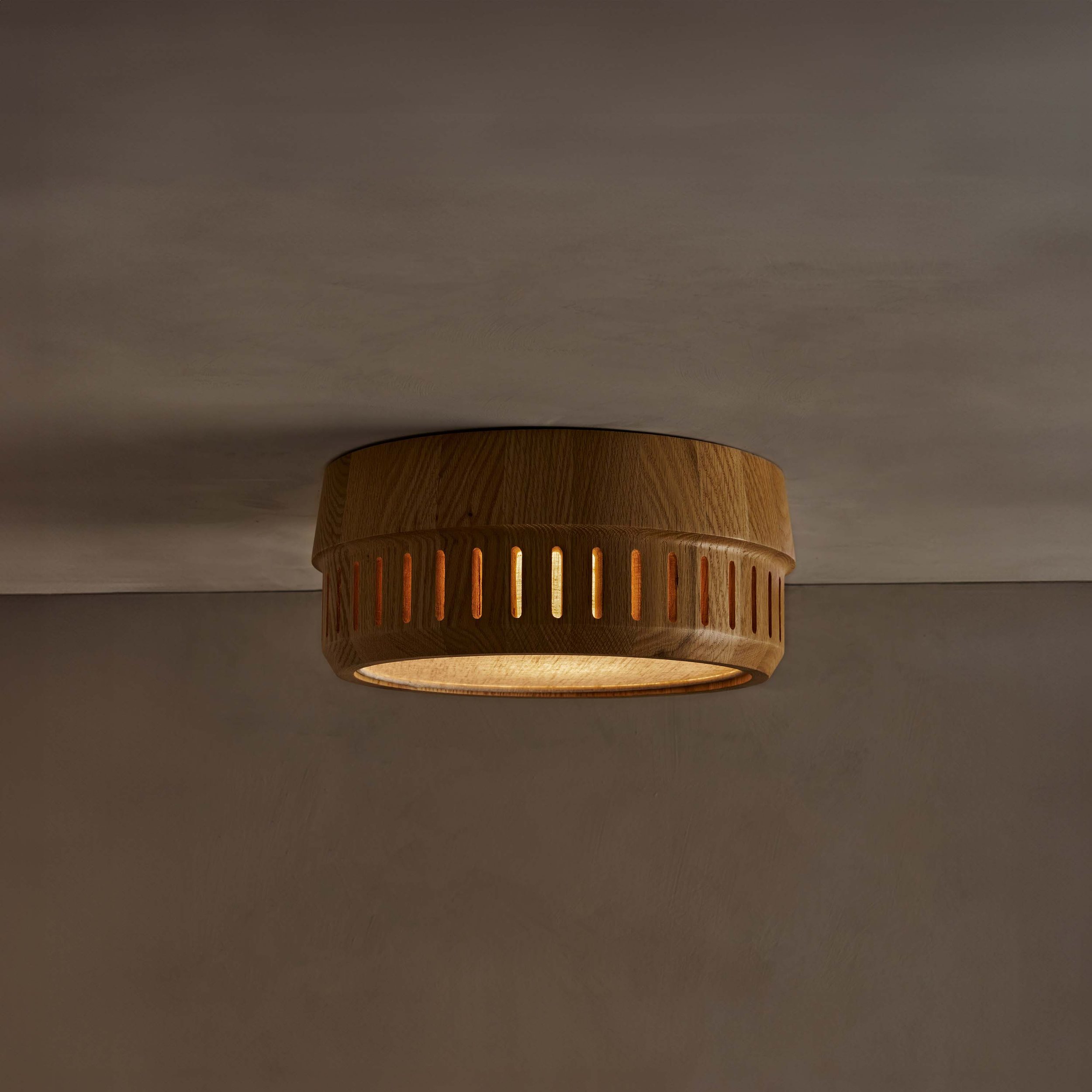 The Adler ceiling mount nests a linen drum shade within a solid wood exterior, offering functional downward illumination. Notches in the wood allow glimpses of the fabric shade within.