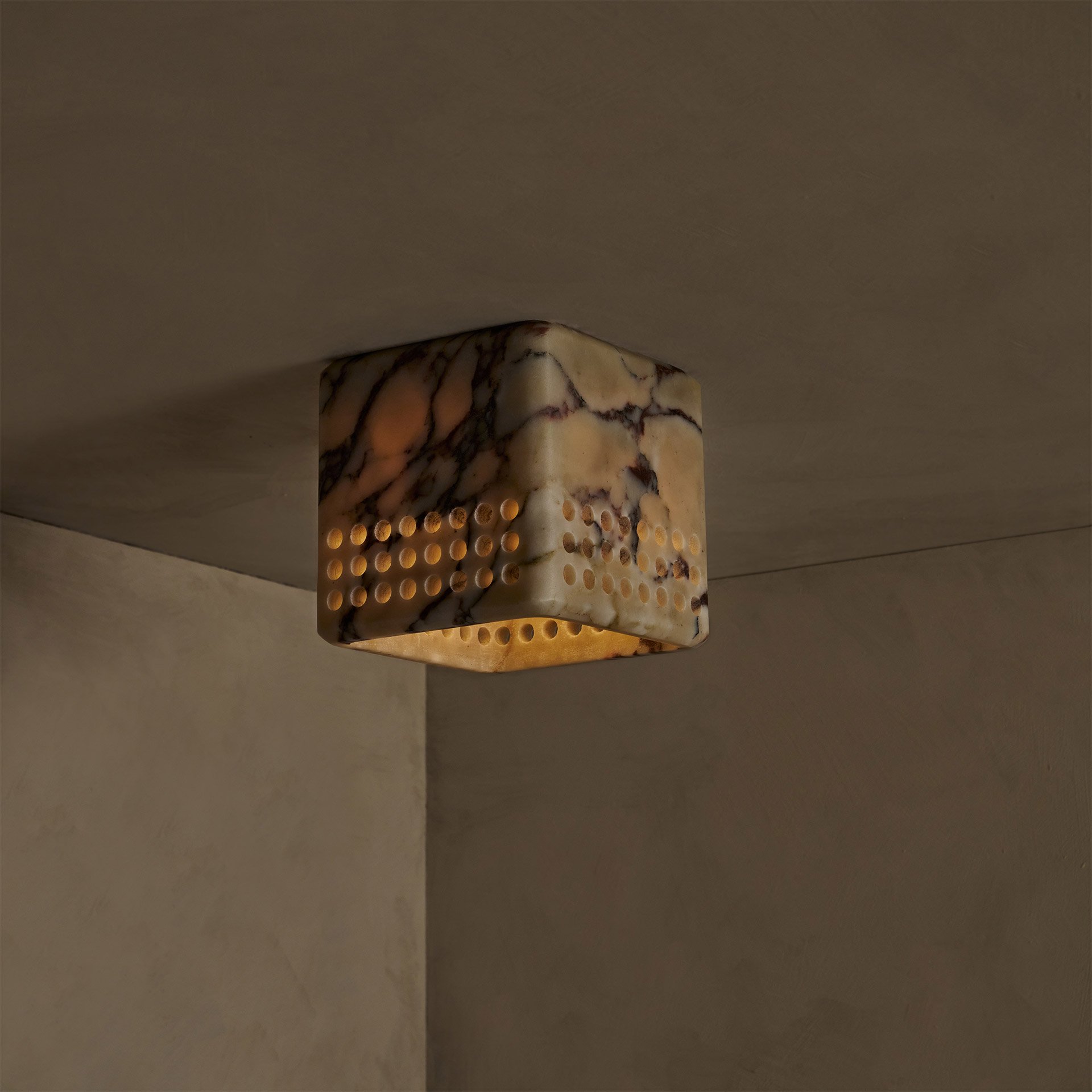 The Bond ceiling mount boasts a simple square form, highlighting its raw stone material. Perforations in the shade emit light downward and outward.