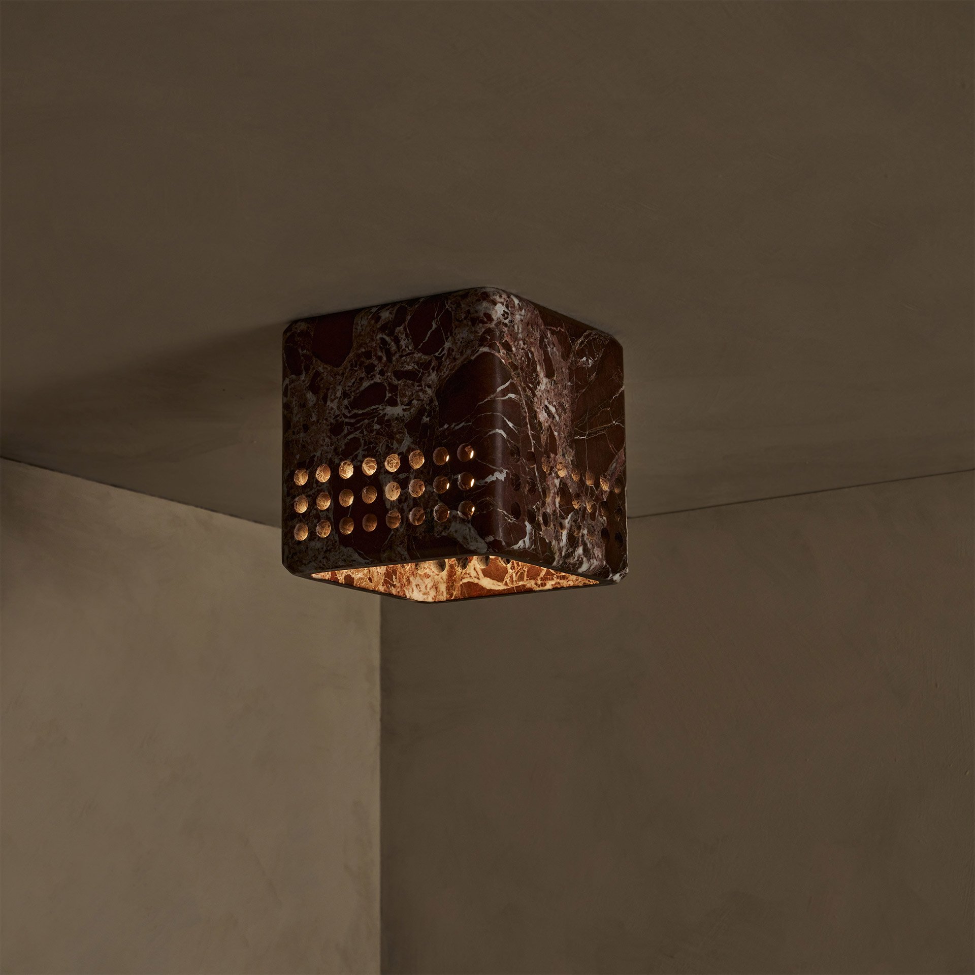 The Bond ceiling mount boasts a simple square form, carved from a solid block, highlighting its raw stone material. Perforations in the shade emit light downward and outward
