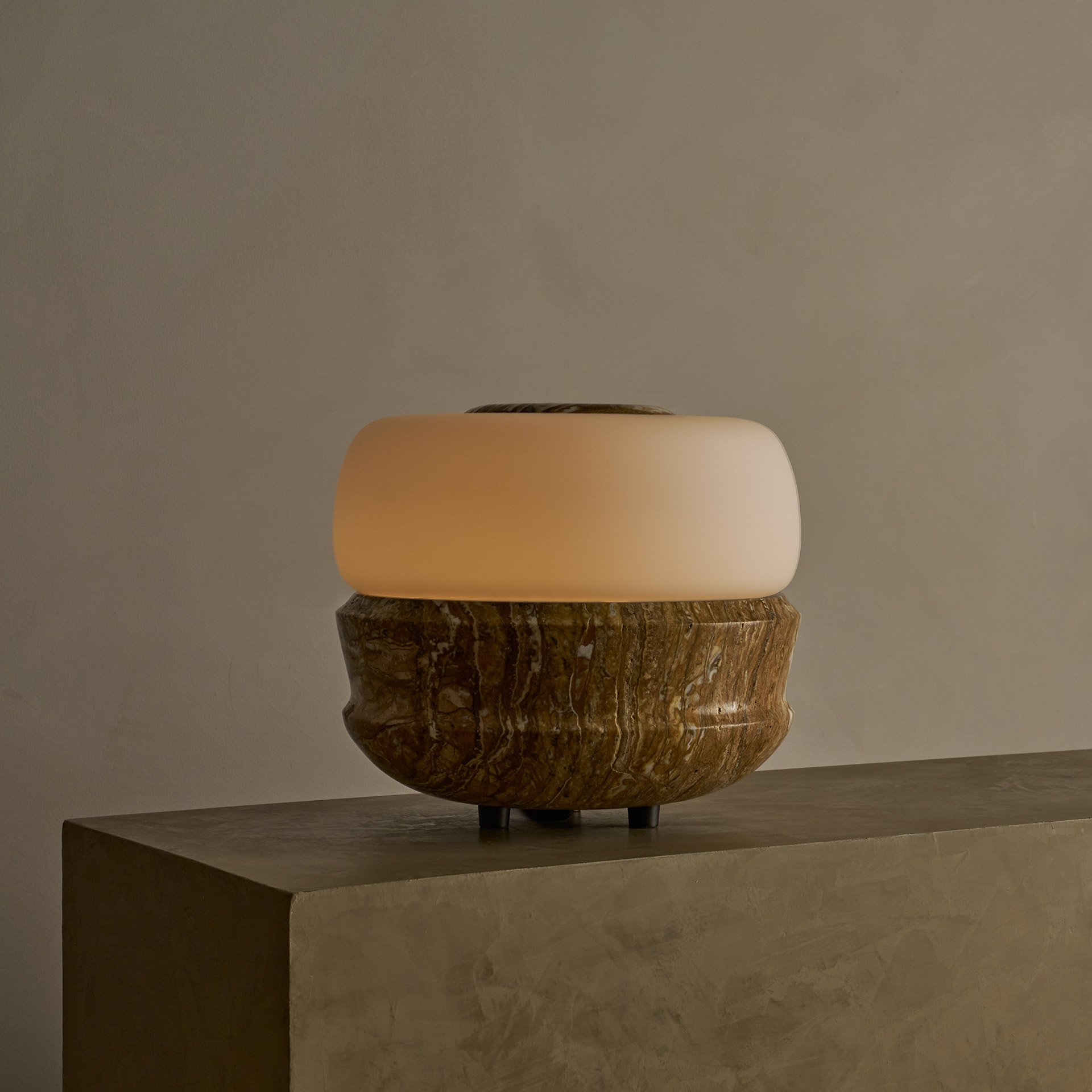The Ramson table lamp boasts a bulbous stone base with intricate geometric details, complemented by a seamless glass shade. Supported by three tapered solid brass feet.