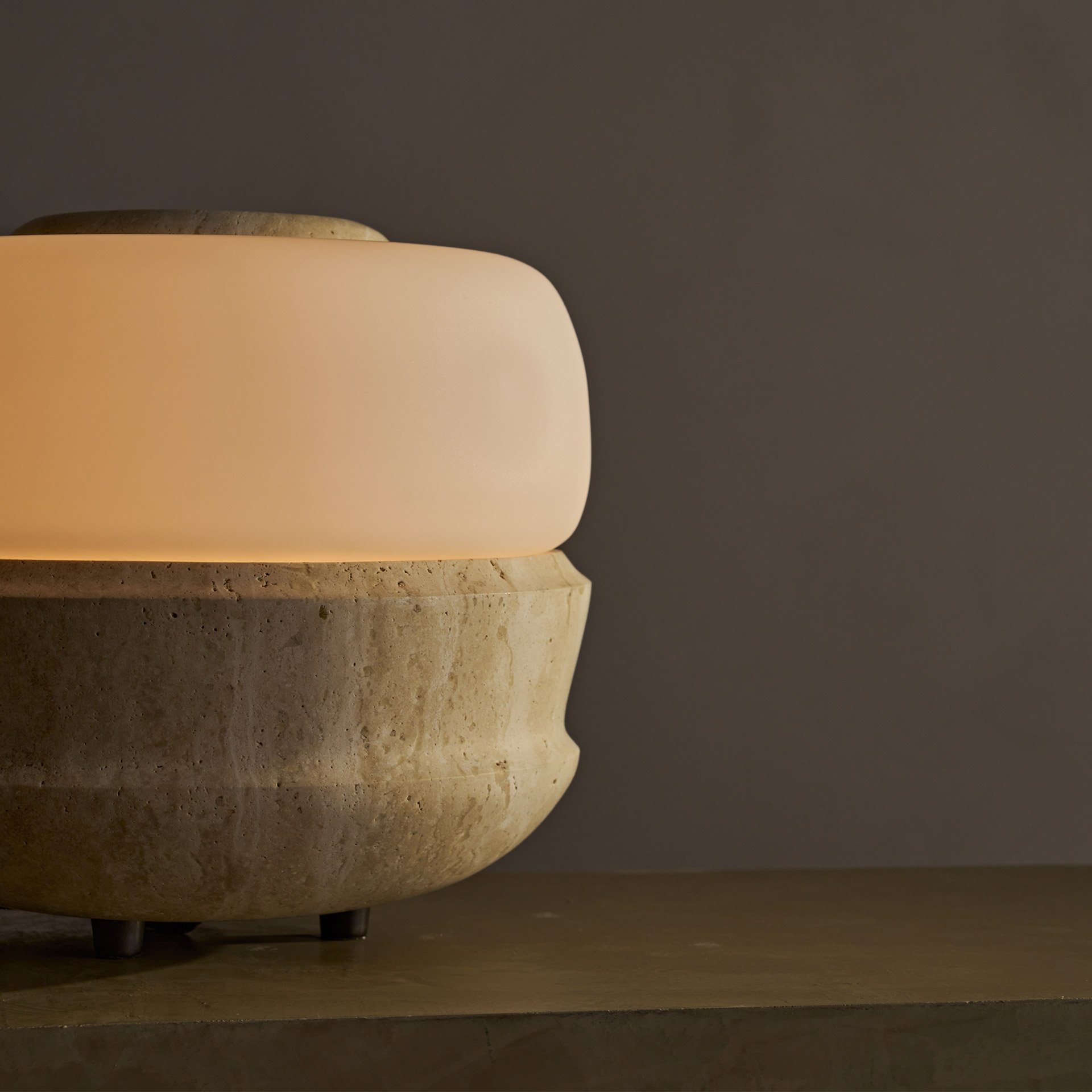 The Ramson table lamp boasts a bulbous stone base with intricate geometric details, complemented by a seamless glass shade. Supported by three tapered solid brass feet.