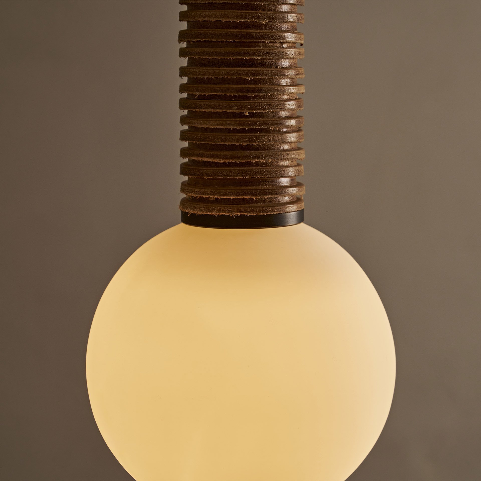 The Pell pendant lamp features a solid body crafted from stacked leather hide disks, showcasing the natural imperfections of the material's edge. A frosted glass sphere hangs at the bottom