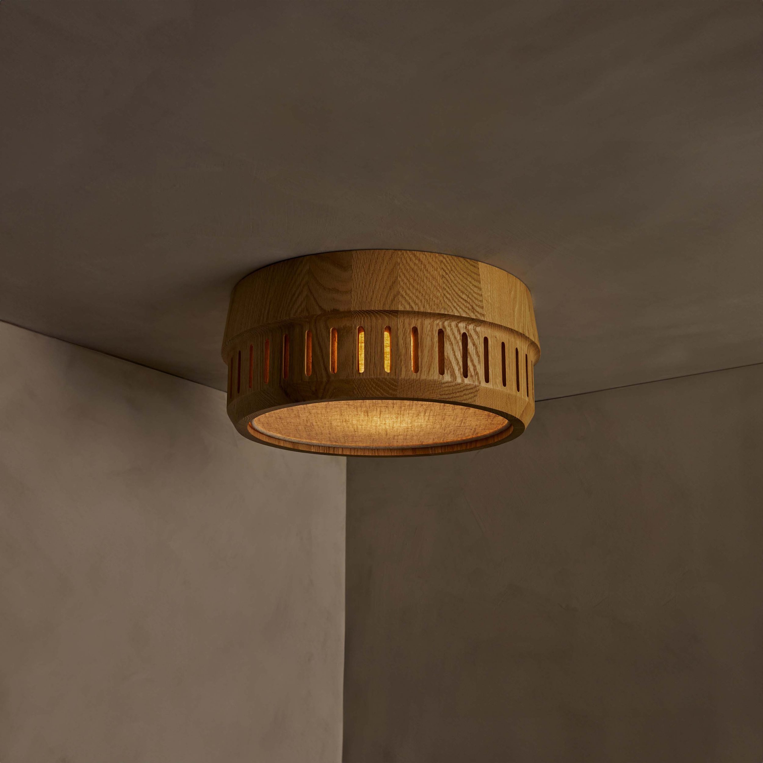 The Adler ceiling mount nests a linen drum shade within a solid wood exterior, offering functional downward illumination. Notches in the wood allow glimpses of the fabric shade within.