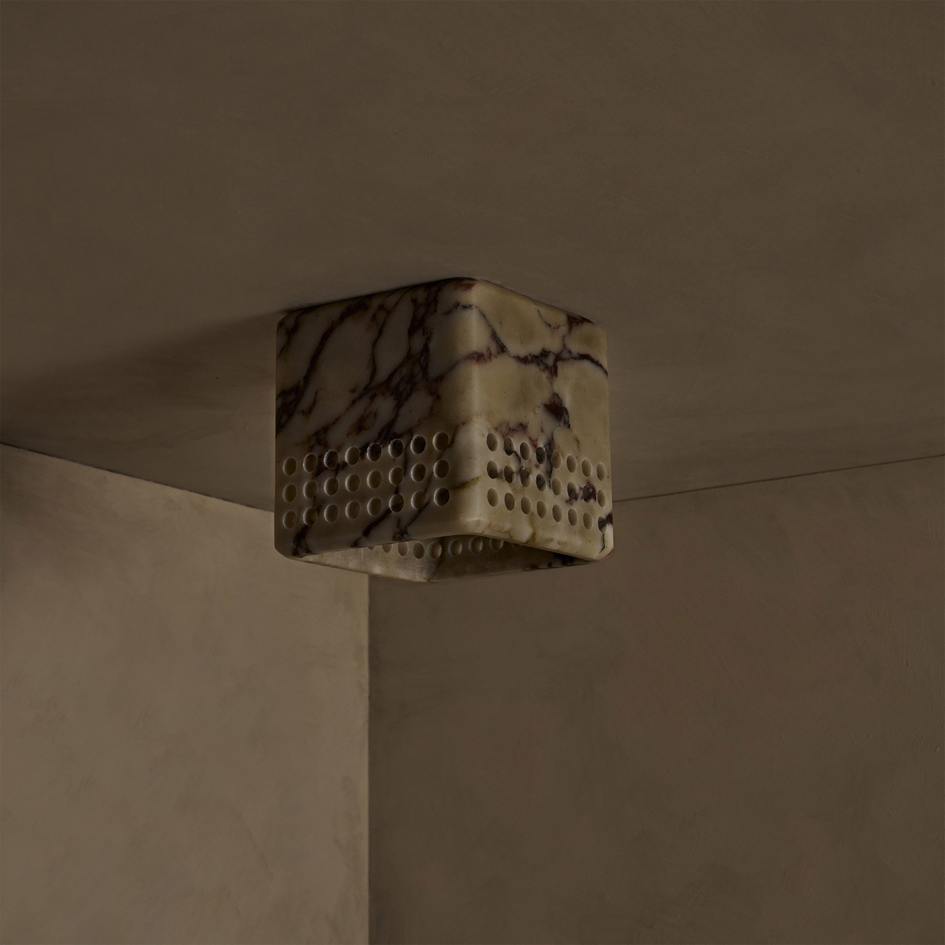 The Bond ceiling mount boasts a simple square form, carved from a solid block, highlighting its raw stone material. Perforations in the shade emit light downward and outward