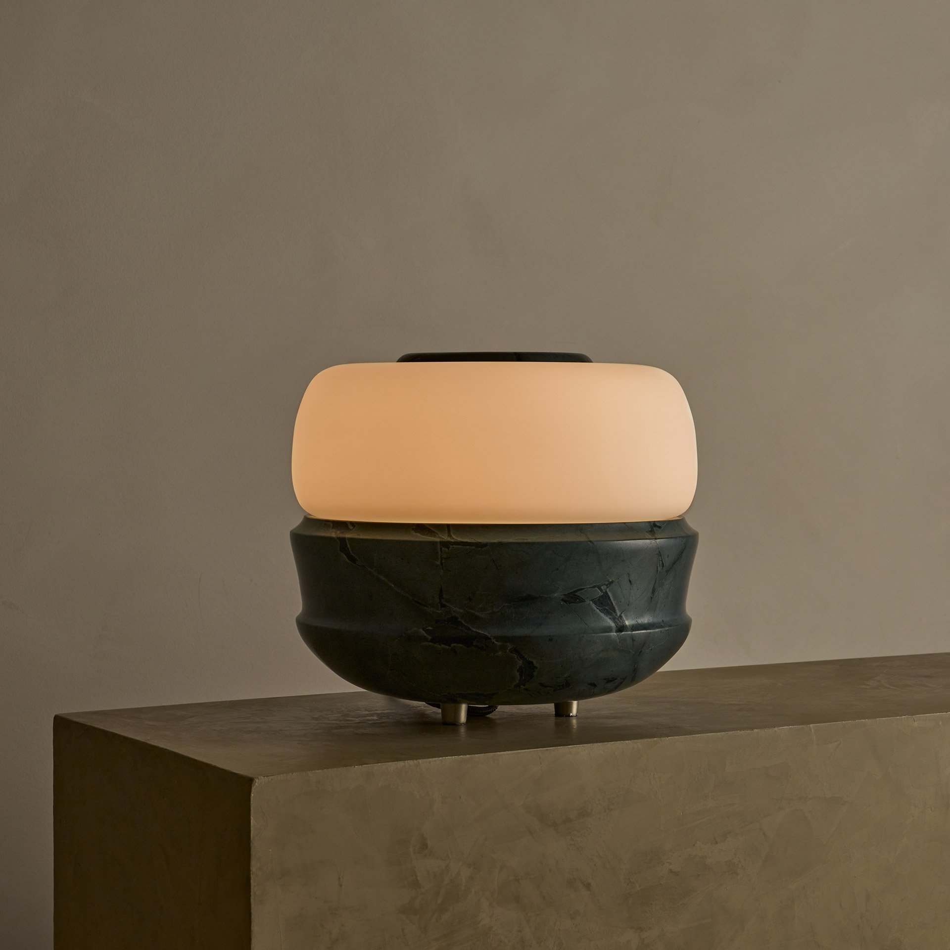 The Ramson table lamp boasts a bulbous stone base with intricate geometric details, complemented by a seamless glass shade. Supported by three tapered solid brass feet.