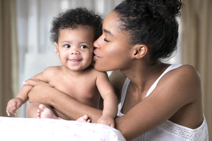 A woman kisses a smiling young child on the cheek inside a home.