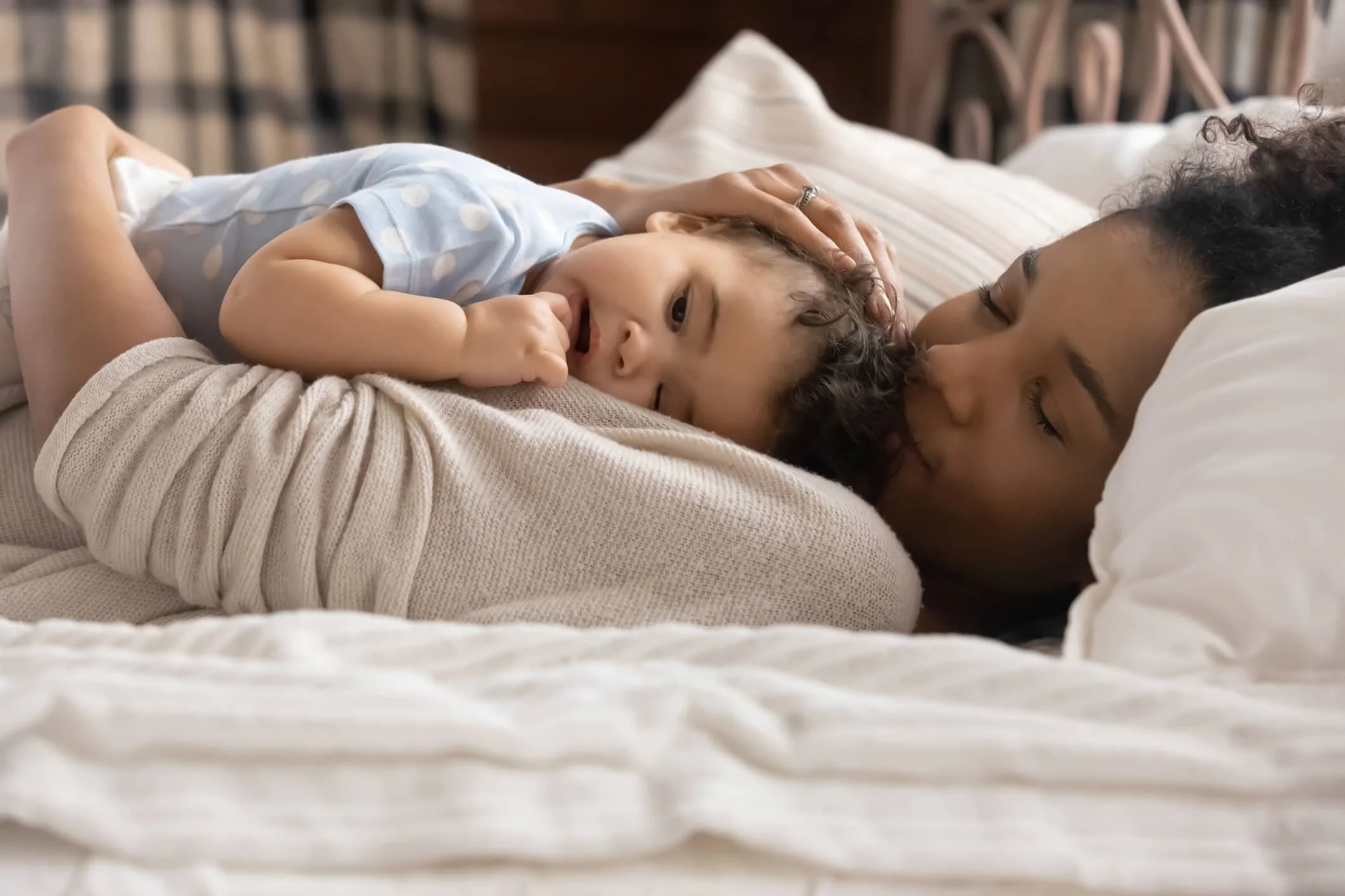 A woman laying in bed cuddling a young child, possibly her son, with her eyes closed and gentle smile, in a cozy bedroom.