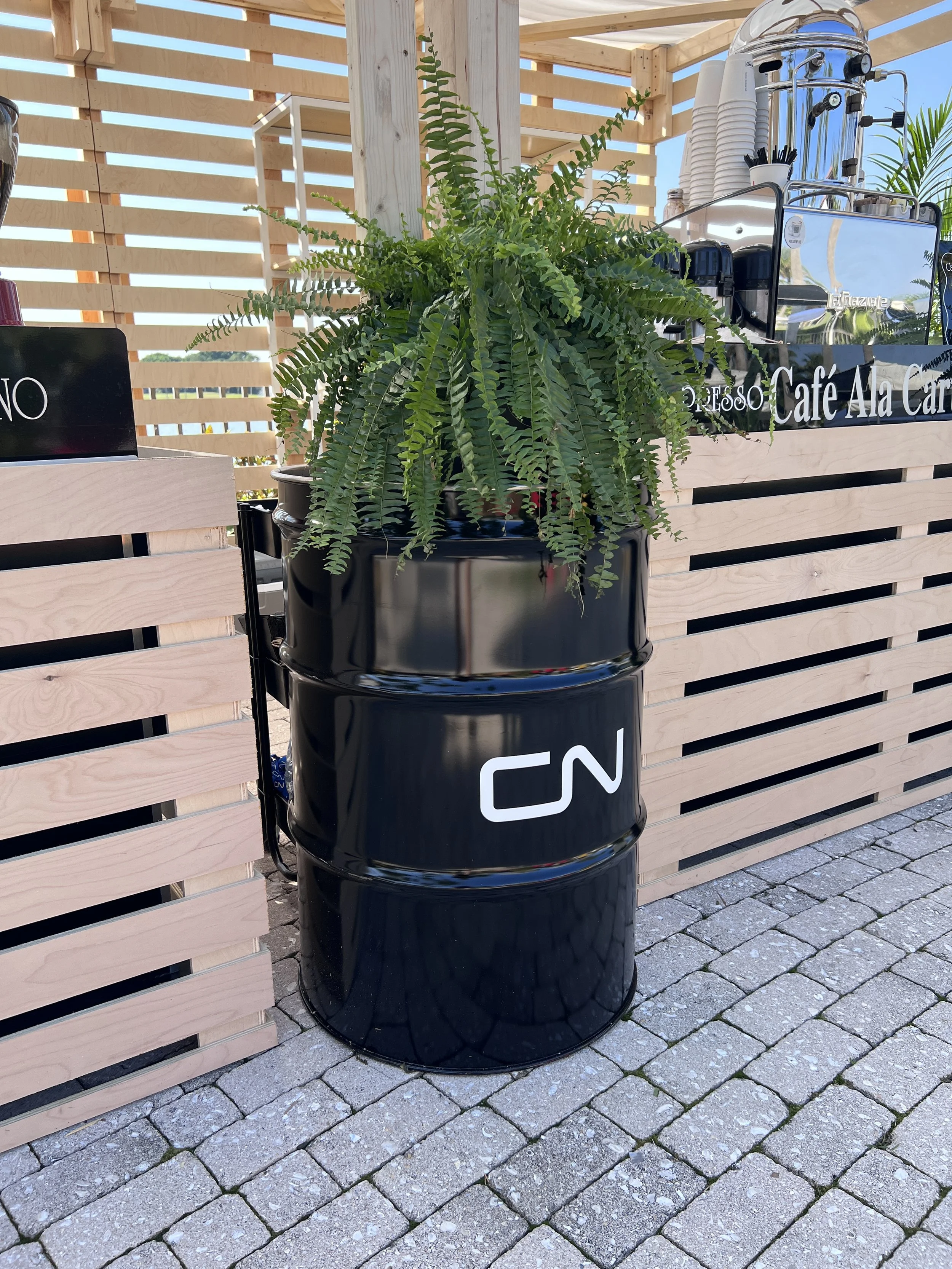 Black barrel planter with a fern and CN branding beside a custom-built coffee cabana