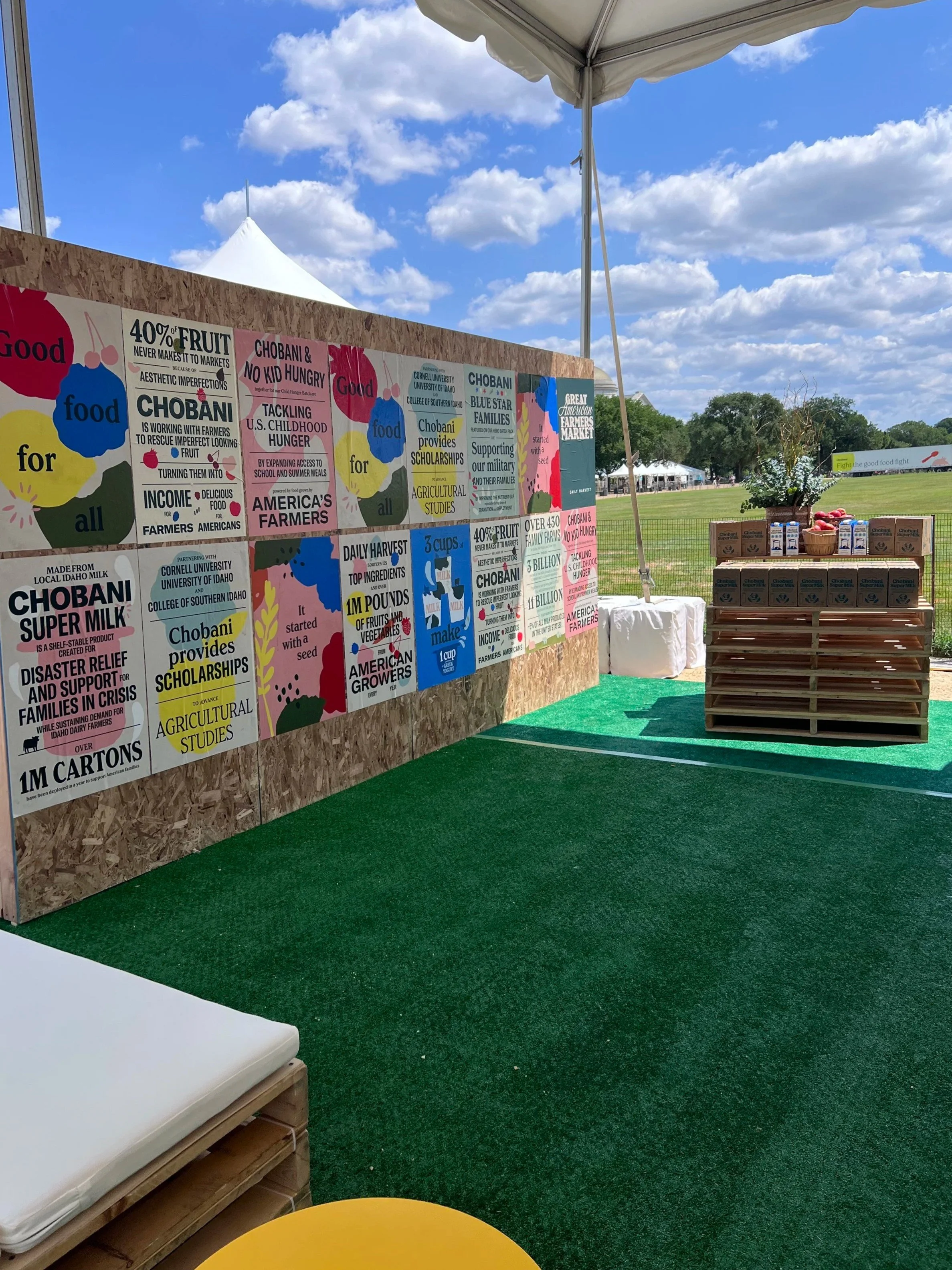Custom-built poster wall with Chobani campaign graphics inside an event tent, with pallet seating on turf