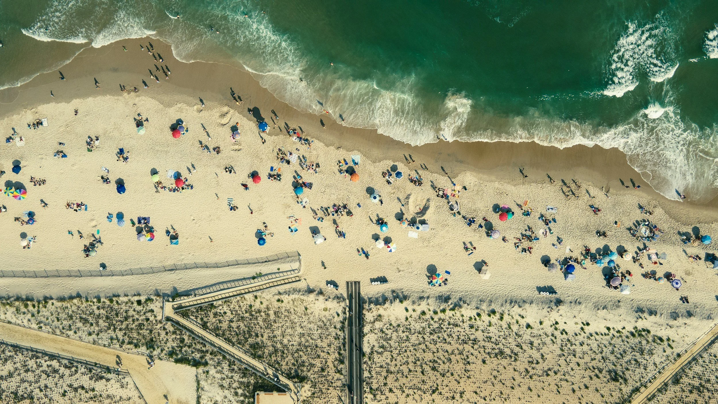 Aerial view of a crowded beach with colorful umbrellas, people walking, and waves crashing on the shore.