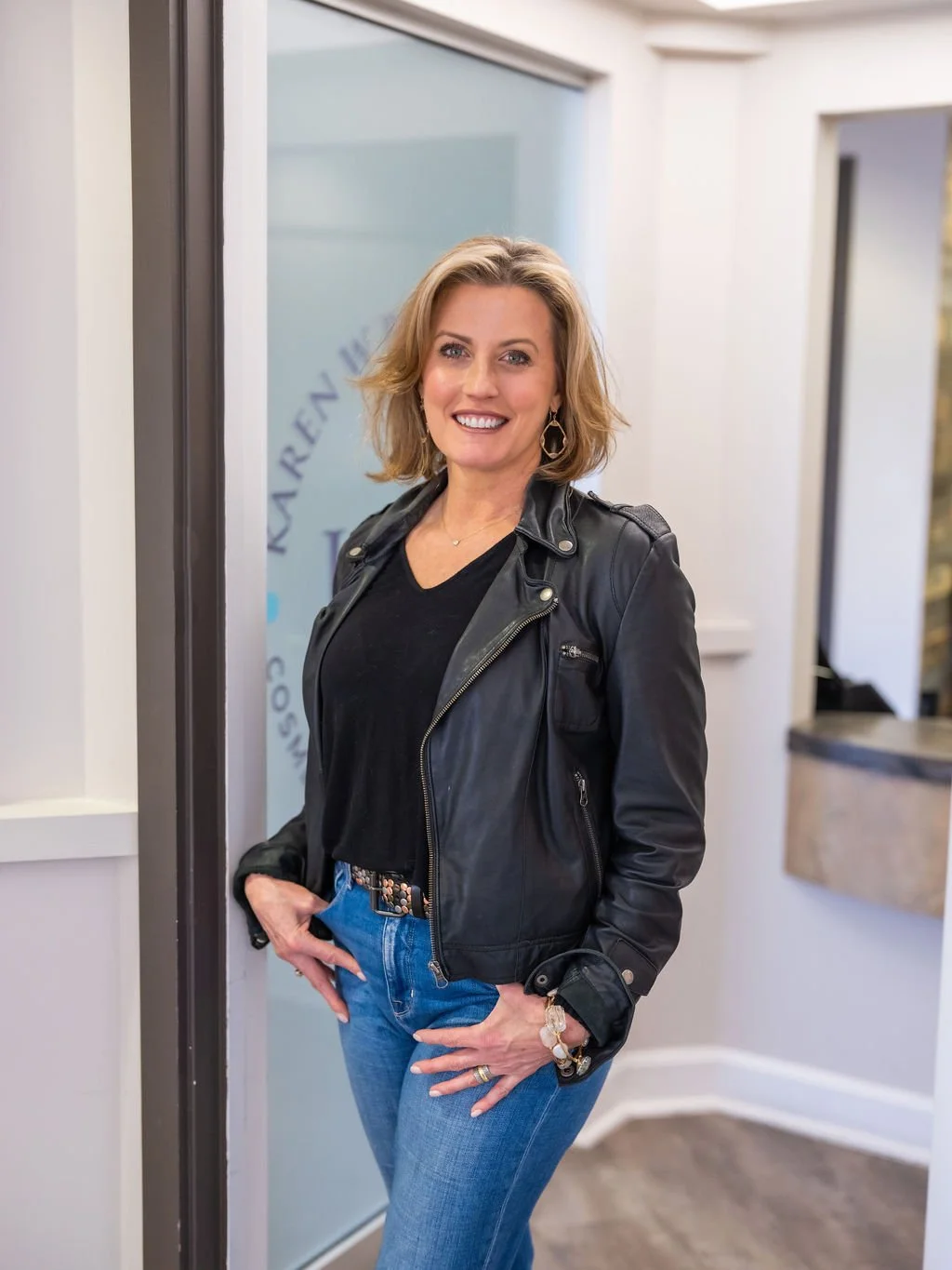 A woman with blonde, shoulder-length hair smiling, wearing a black leather jacket, black shirt, and blue jeans, standing indoors near a glass door.