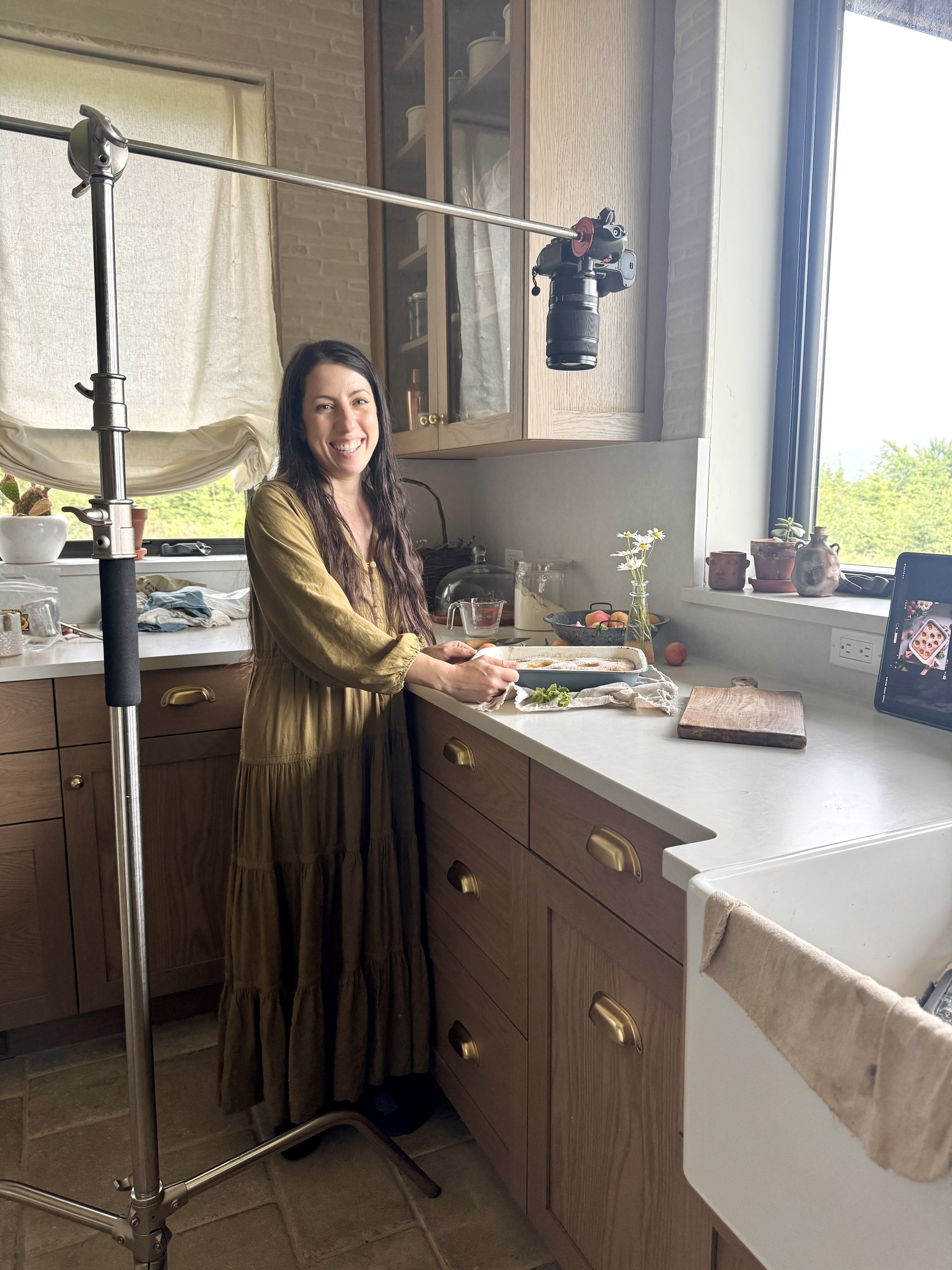 Eva Kosmas Flores smiling in her custom homestead kitchen with gold hardware and natural light, standing beside a camera rig and a freshly baked apricot cake