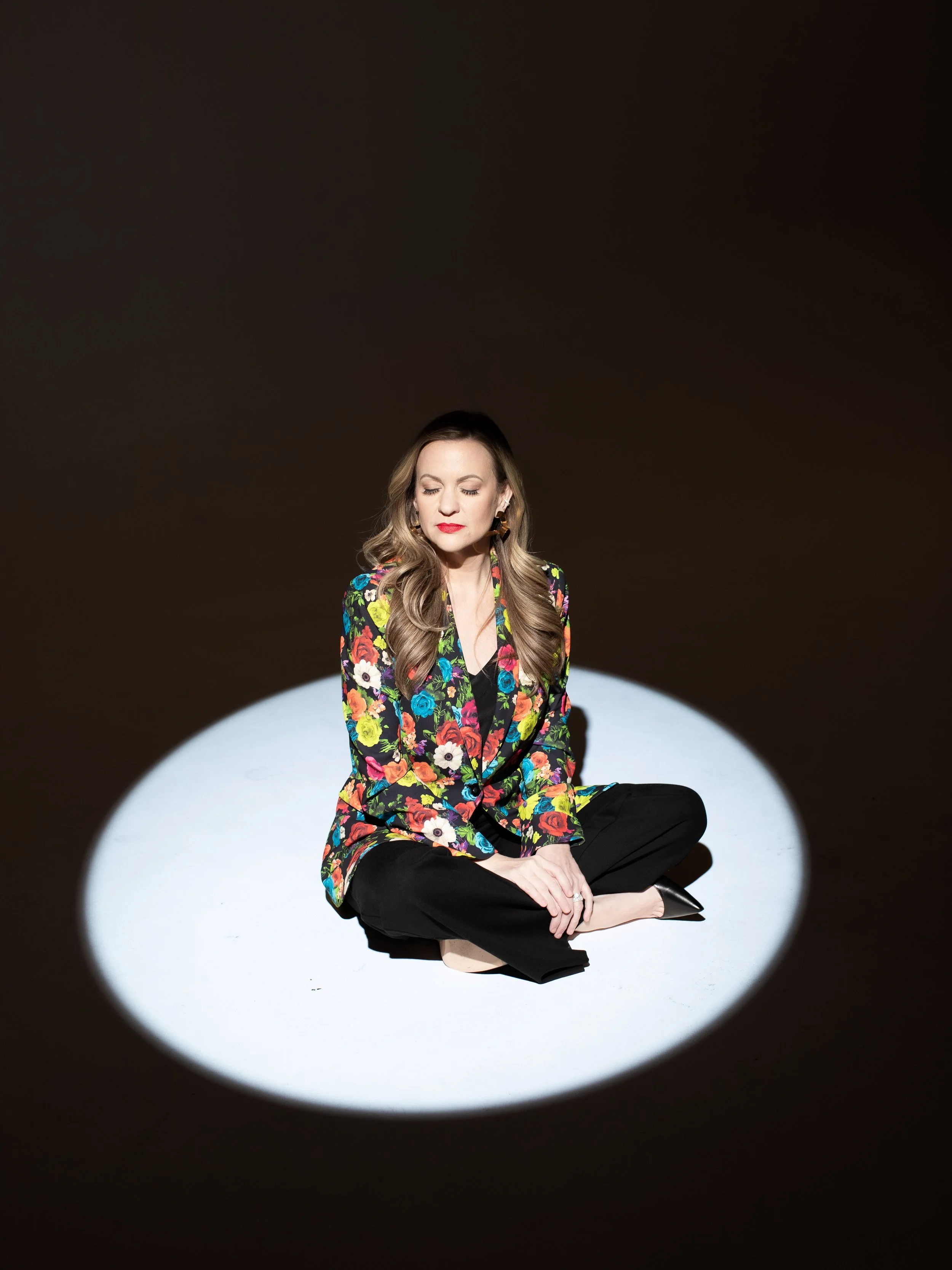 woman sitting in spotlight wearing a floral blazer and eyes closed