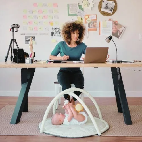 A woman working on a laptop. A baby is playing on the floor next to her.