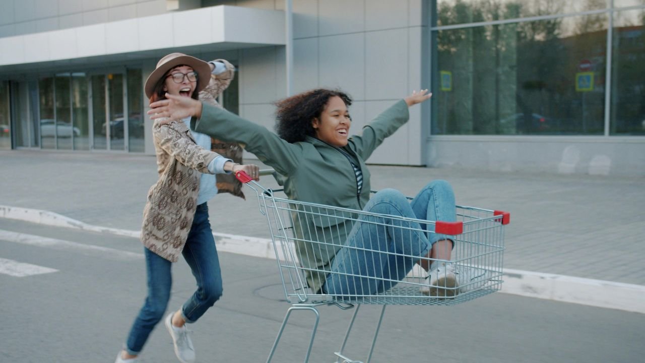 One person pushing another person down the street in a shopping cart.