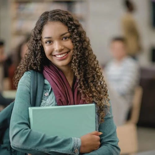 A young person standing in a classroom, smiling, and holding a binder.