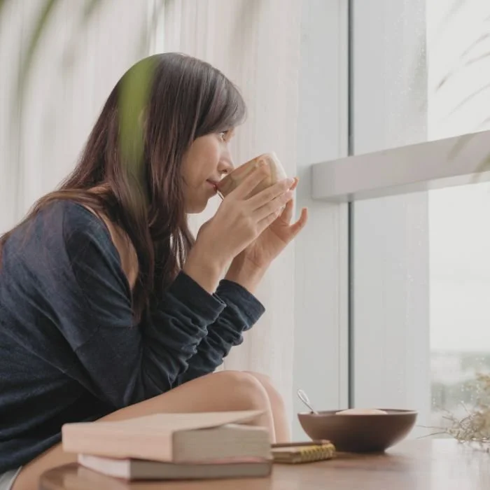 Person sipping coffee while having her breakfast. She is looking out a window and there are books on the table beside her.