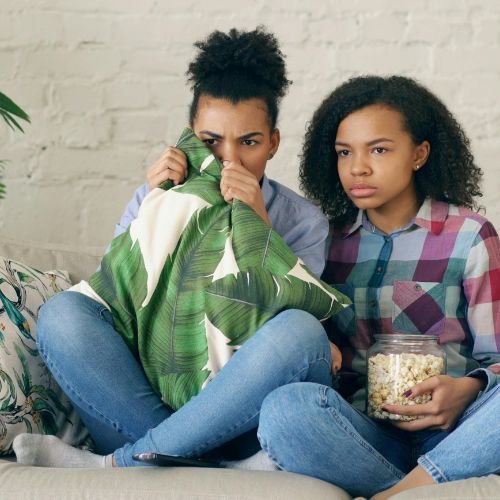 Two people sitting on a sofa, watching a movie. One is holding popcorn.