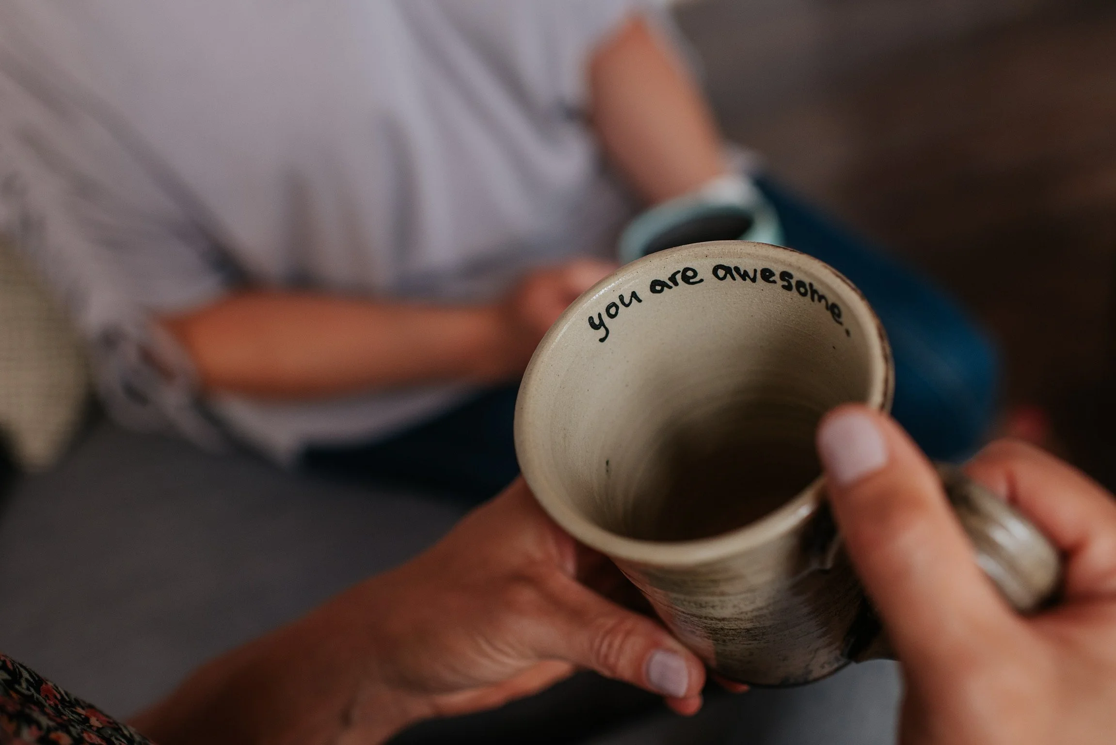 A person holding a beige mug with the words "you are awesome" written inside, with another person sitting nearby.
