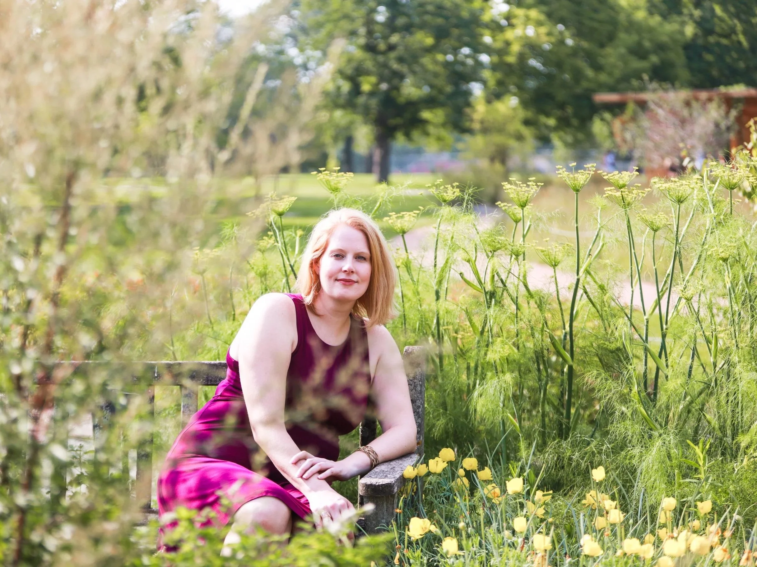Woman with red hair wearing a purple dress sitting on a bench in a garden with yellow flowers and greenery in the background.