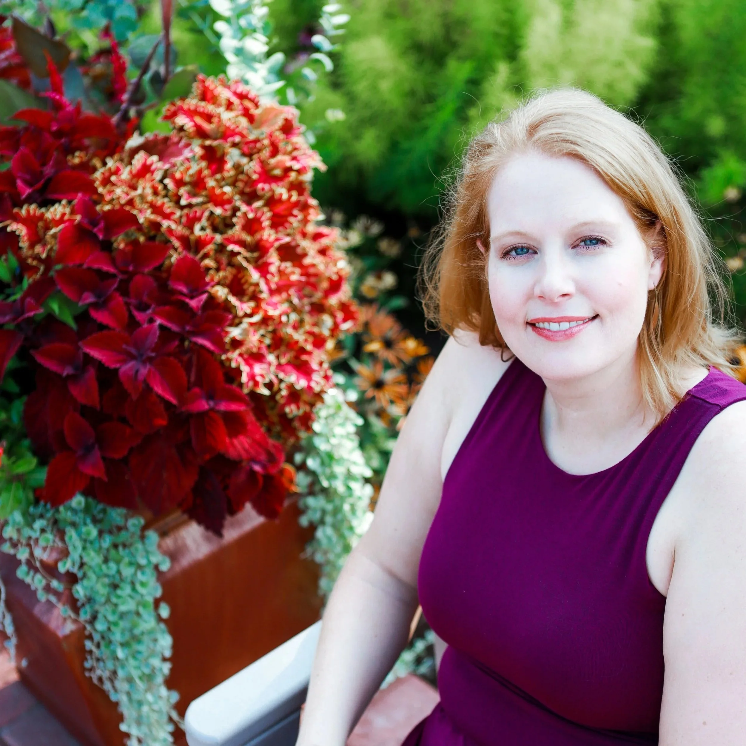 A woman with light skin and blue eyes wearing a purple sleeveless dress standing outdoors next to red and green plants with a brick border.