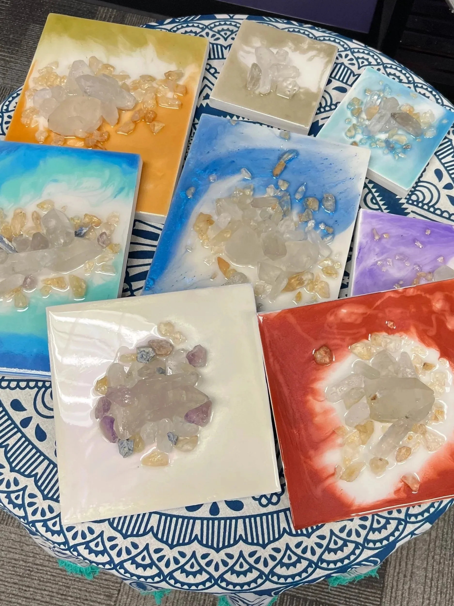 Several painted canvases with abstract colorful backgrounds and crystals on top, arranged on a round table with a blue and white patterned tablecloth.
