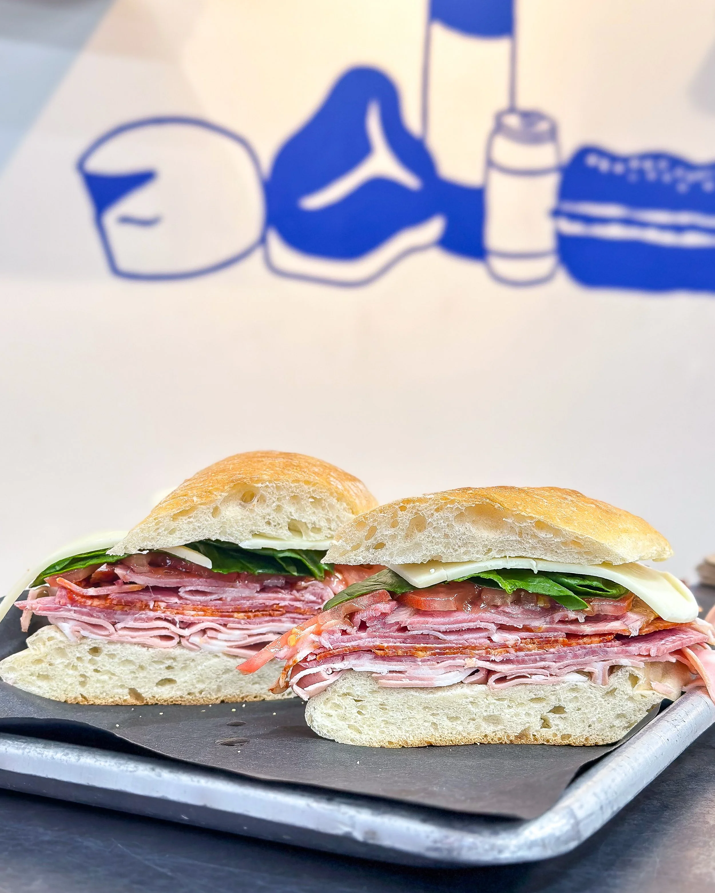 A sandwich with sliced ham, lettuce, tomato, and cheese on a baguette, served on a black slate platter with a metal utensil in the foreground.