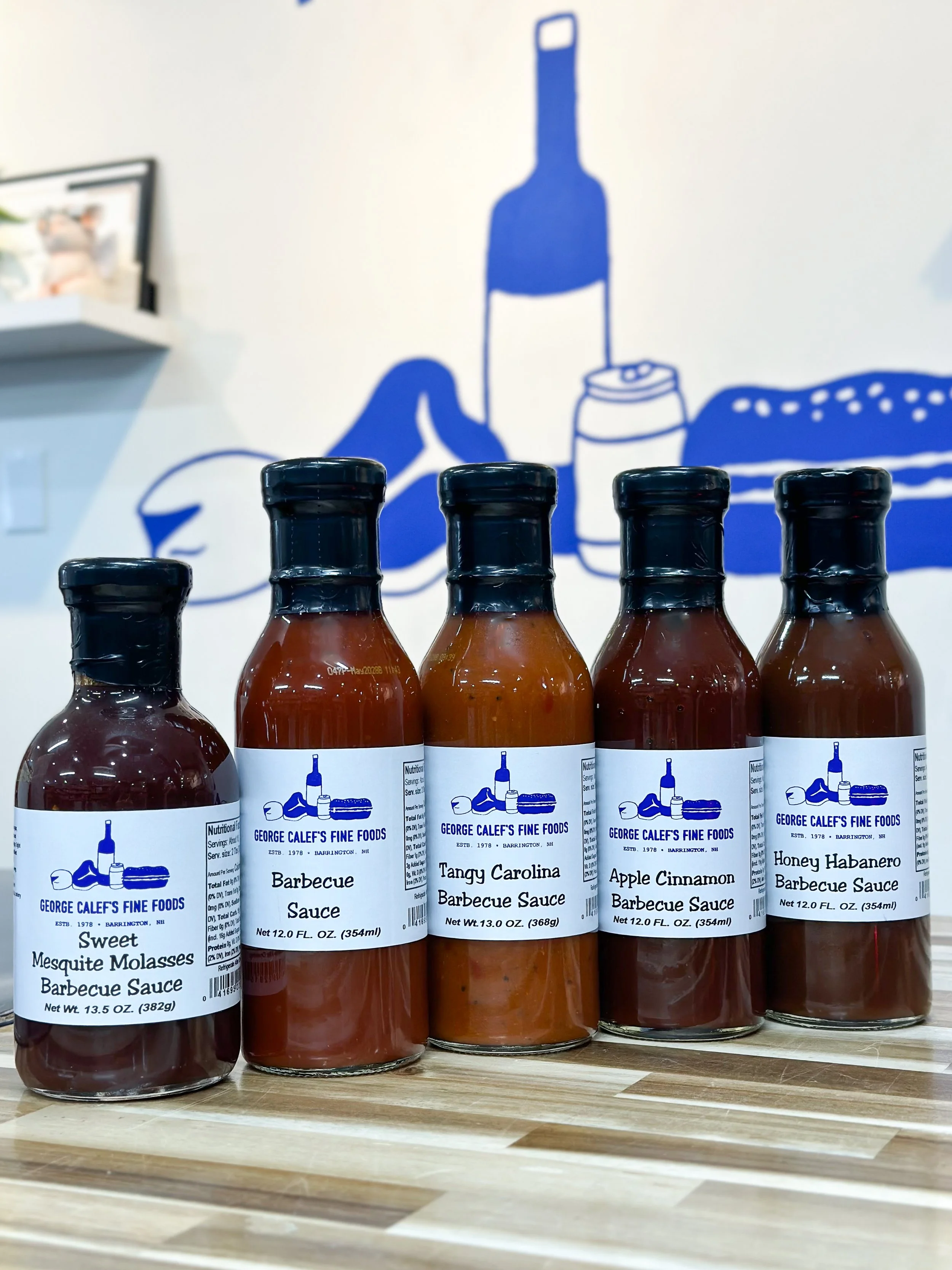 A row of four bottles of barbecue sauce on a wooden surface, labeled as George Calef's Fine Foods, with three different flavors: Sweet Mesquite Molasses, Tangy Carolina, and Apple Cinnamon. A blue and white mural of a beer bottle, hamburger, and soda can is visible in the background.