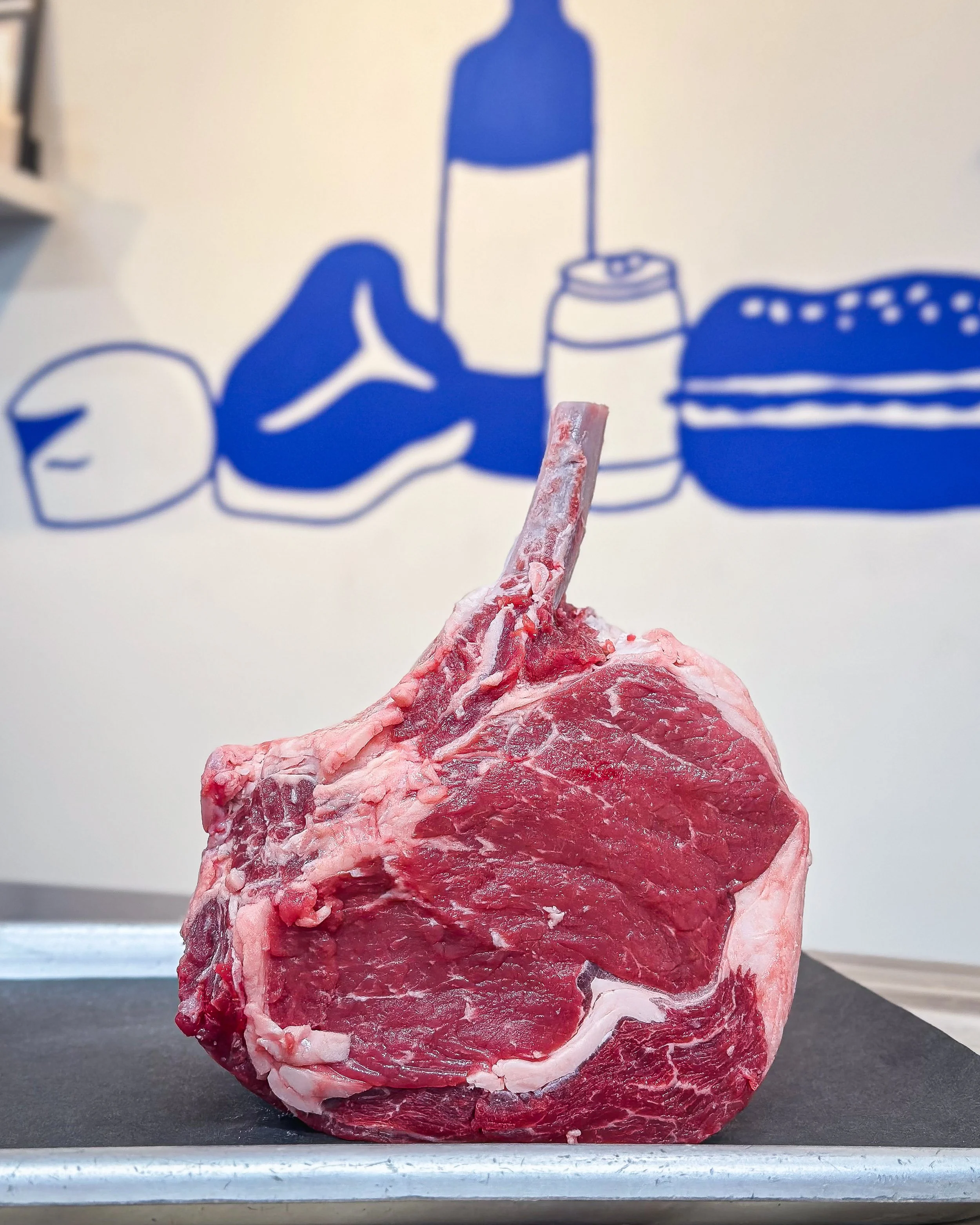 A raw piece of meat with a bone sticking out, placed on a dark surface with a blue and white background featuring illustrations of a fish, a bottle, a can, and a stack of what appears to be plates or food containers.