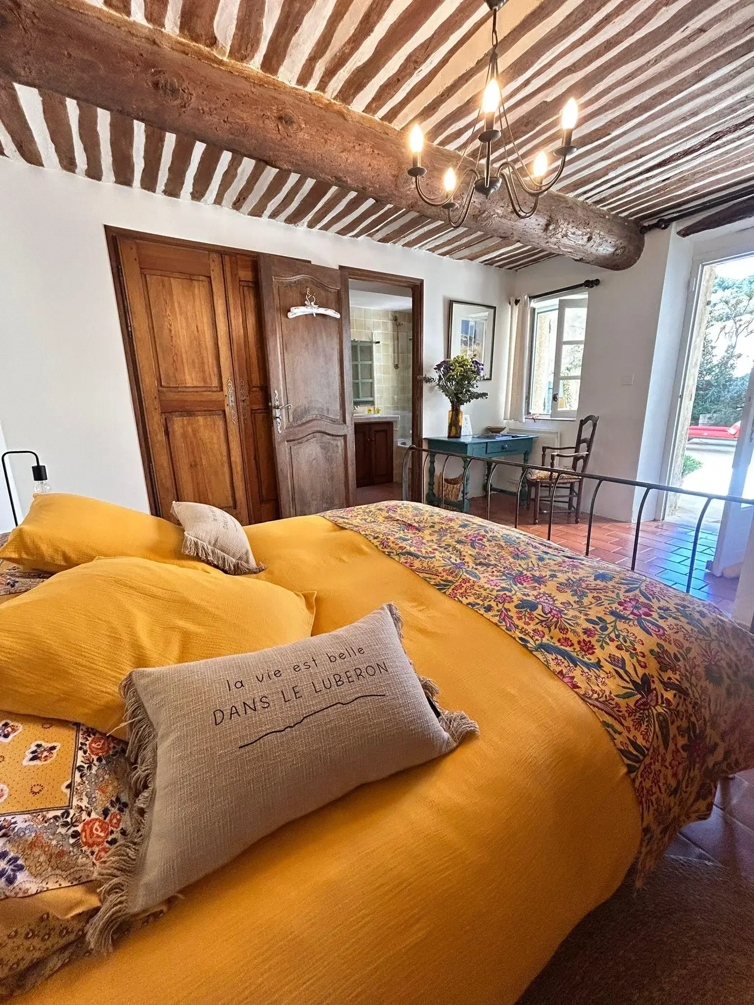 Lit avec des draps jaunes, un coussin avec une phrase en français, une couverture à motifs colorés, dans une chambre avec une poutre en bois au plafond, un lustre en fer, une table avec une vase de fleurs, une fenêtre ouverte et une porte menant à un
